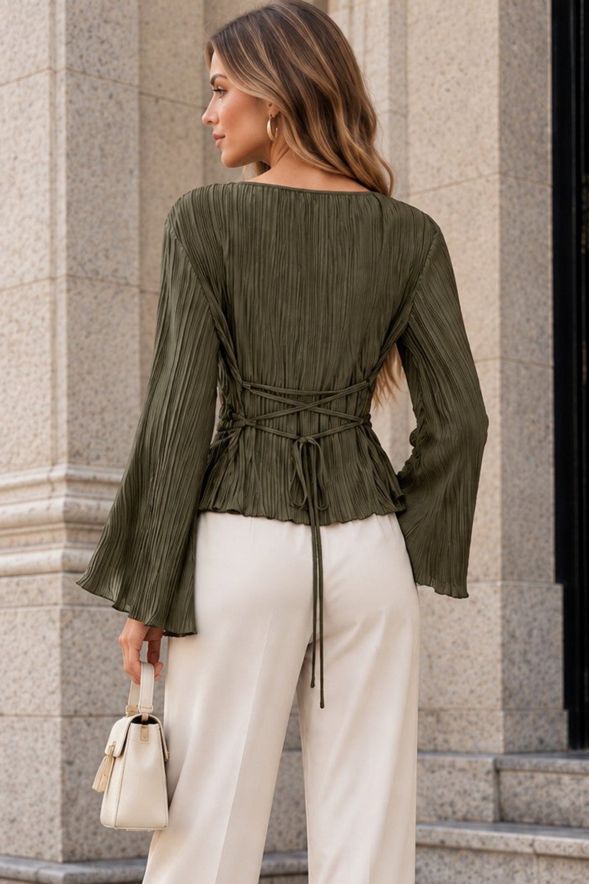 A woman stands outdoors in front of a stone building, shown from the back wearing an olive green pleated long-sleeve top with a tie waist and cream wide-leg pants.
