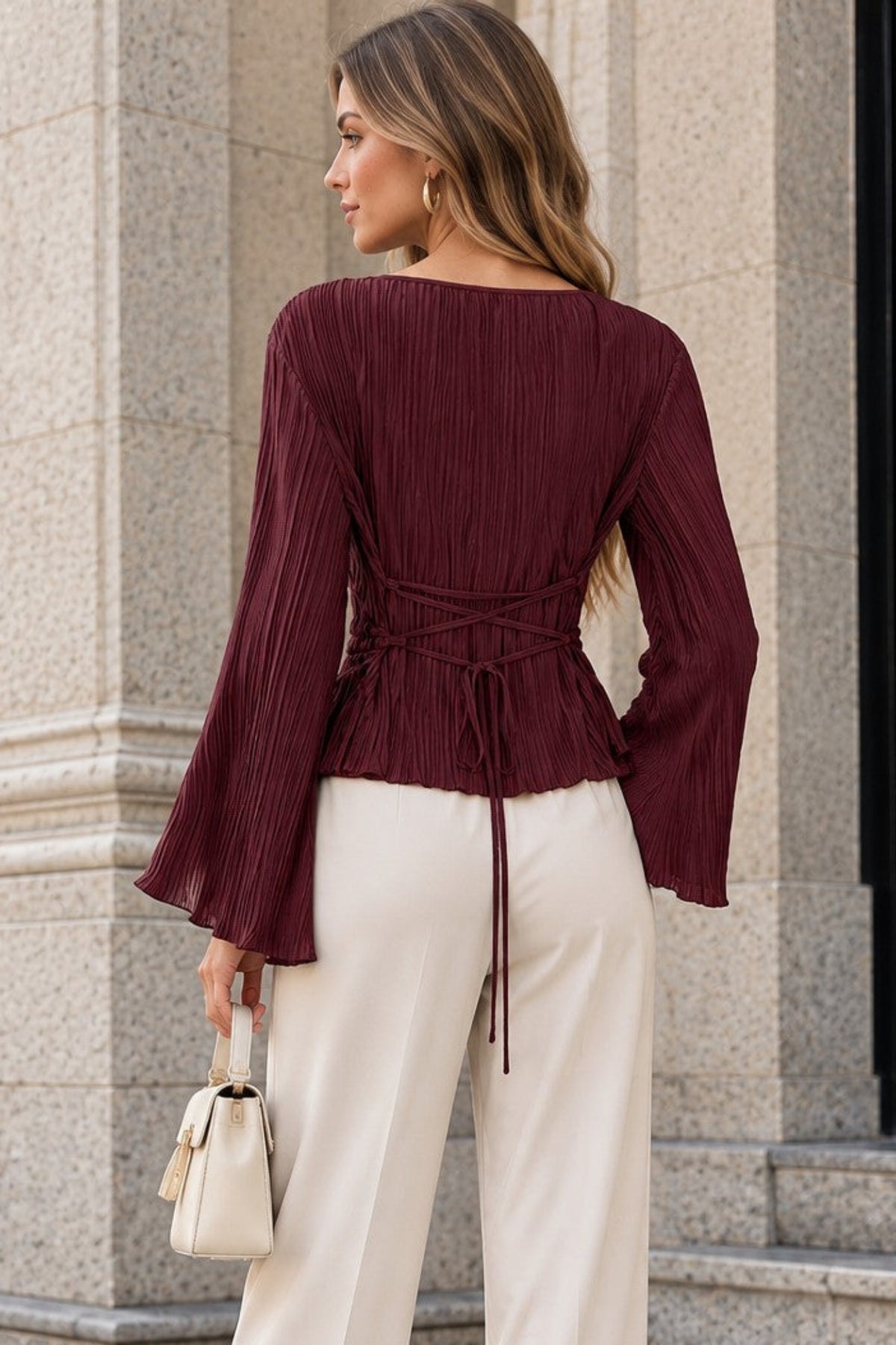 A woman stands outdoors in front of a stone building, shown from the back. She is wearing a deep burgundy pleated long-sleeve top with a tied waist and matching tie detail, paired with cream wide-leg pants and holding a small cream handbag.