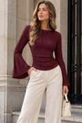 A woman stands outdoors in front of a stone building, wearing a fitted long-sleeve burgundy top with dramatic flared bell sleeves and high-waisted off-white wide-leg pants. She is holding a small beige handbag.