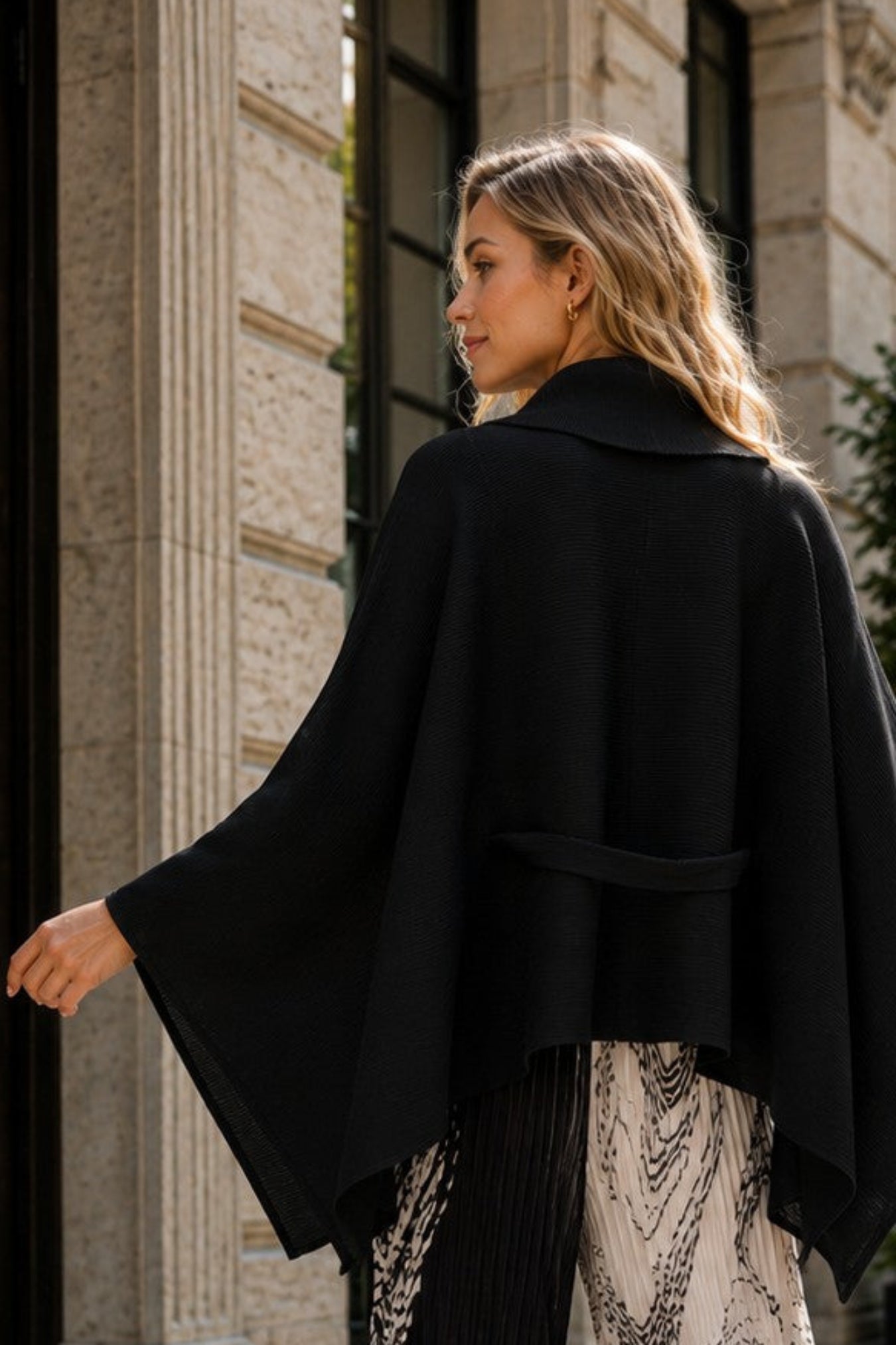 A woman stands on a sunlit cobblestone street in front of a stone building, wearing a black draped cape-style top and wide-leg pleated pants with a black-and-white abstract pattern.