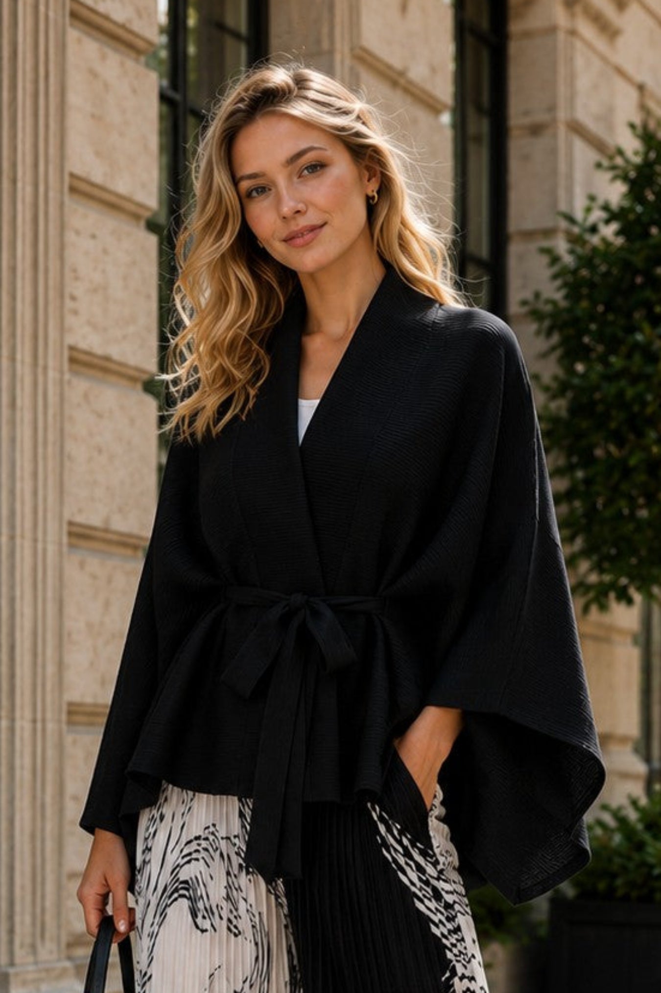 A woman stands on a cobblestone street wearing a black wrap-style jacket and wide-leg pleated trousers with a black-and-white abstract print. The background shows a stone building facade with windows and potted greenery.