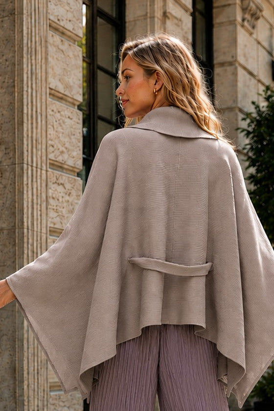 A woman stands on a cobblestone street wearing a light taupe draped cape-style top and lavender pleated wide-leg trousers, photographed outdoors in front of a stone building.