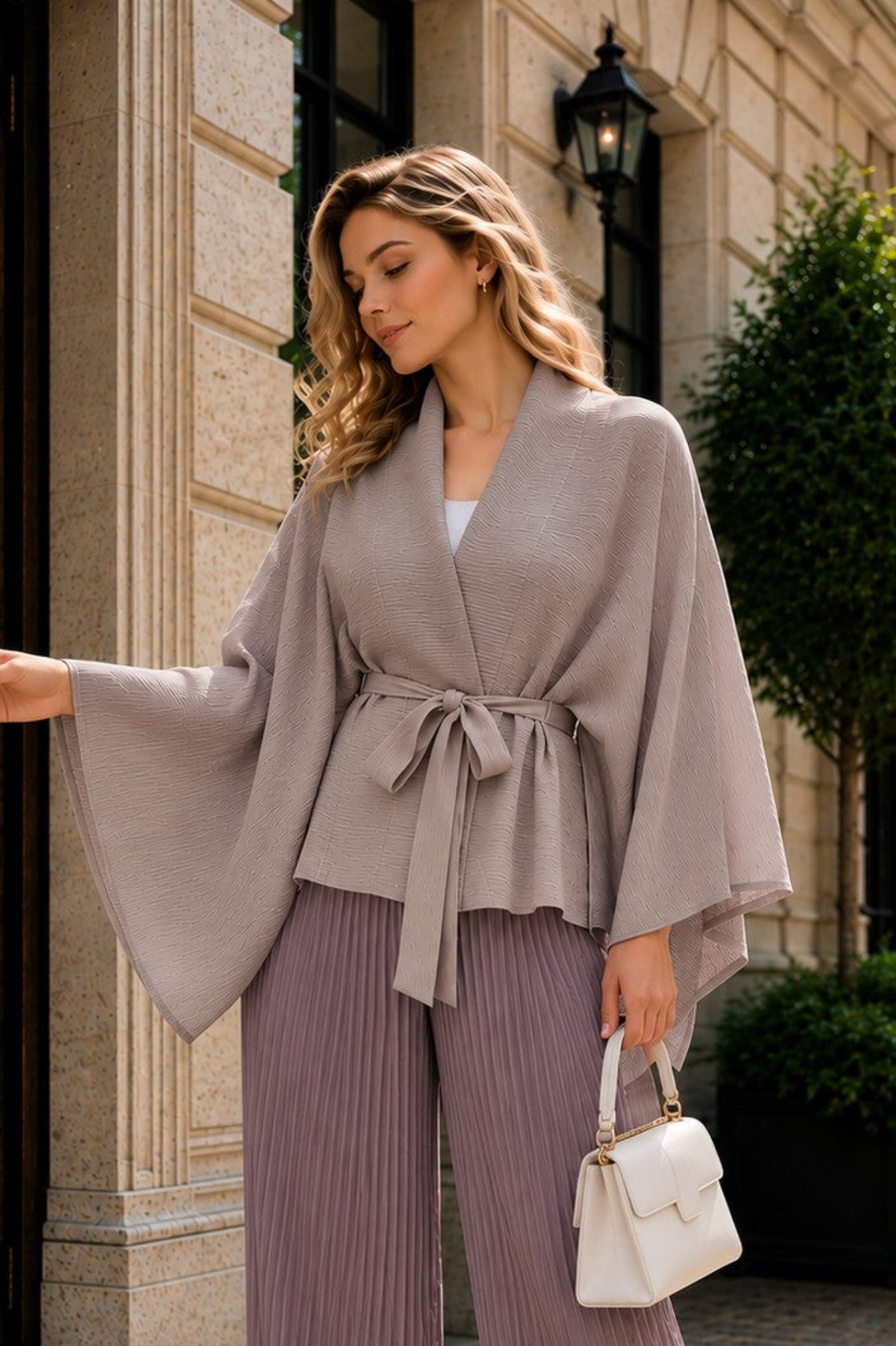 A woman stands on a cobblestone street in front of a beige stone building, wearing a taupe wrap cardigan with wide bell sleeves and matching pleated lavender wide-leg pants, holding a small white handbag.