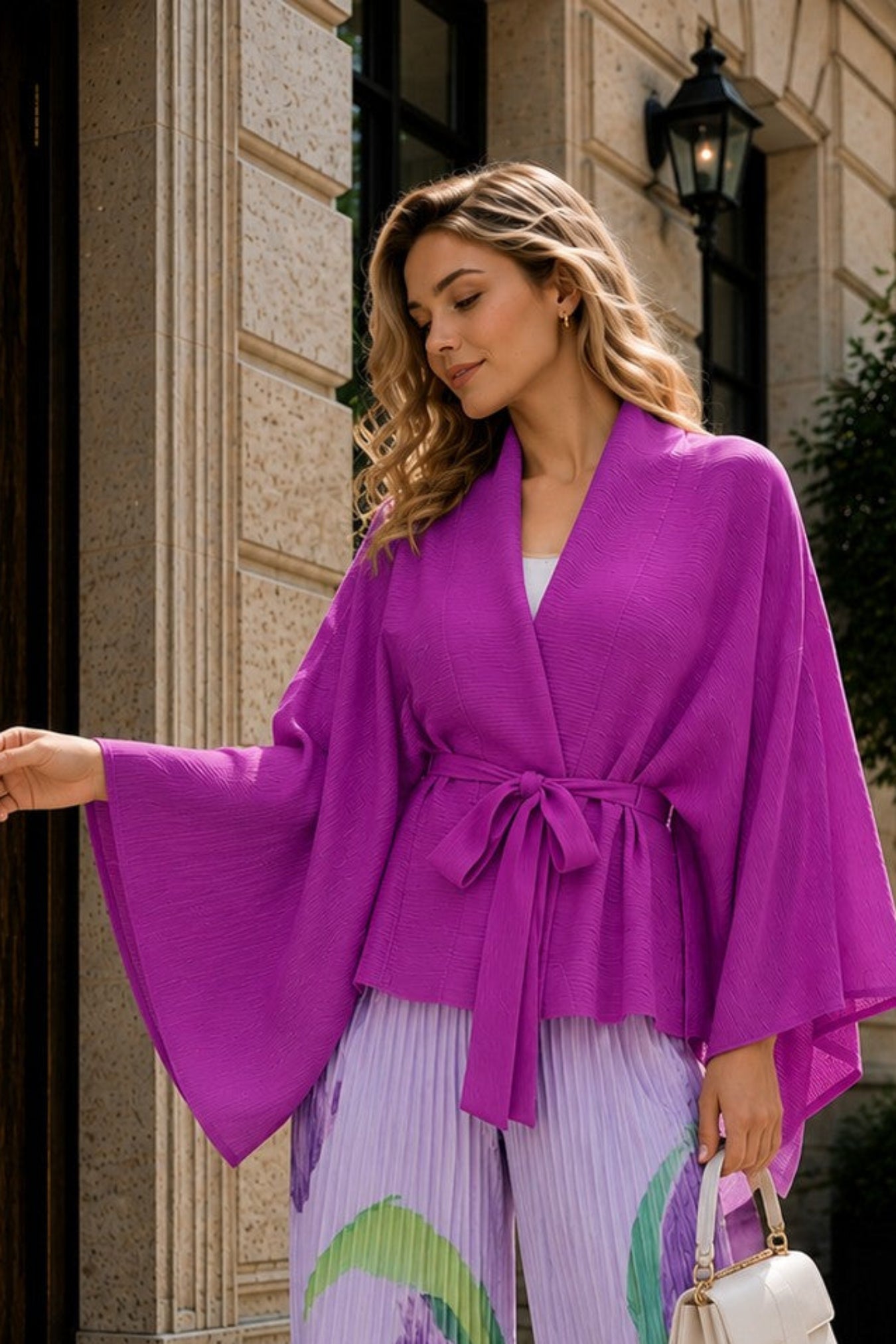 A woman stands on a cobblestone street in front of a stone building, wearing a bright purple wrap-style top with wide bell sleeves and matching pleated wide-leg pants in lavender with abstract green and purple brushstroke patterns. She is holding a small white handbag.