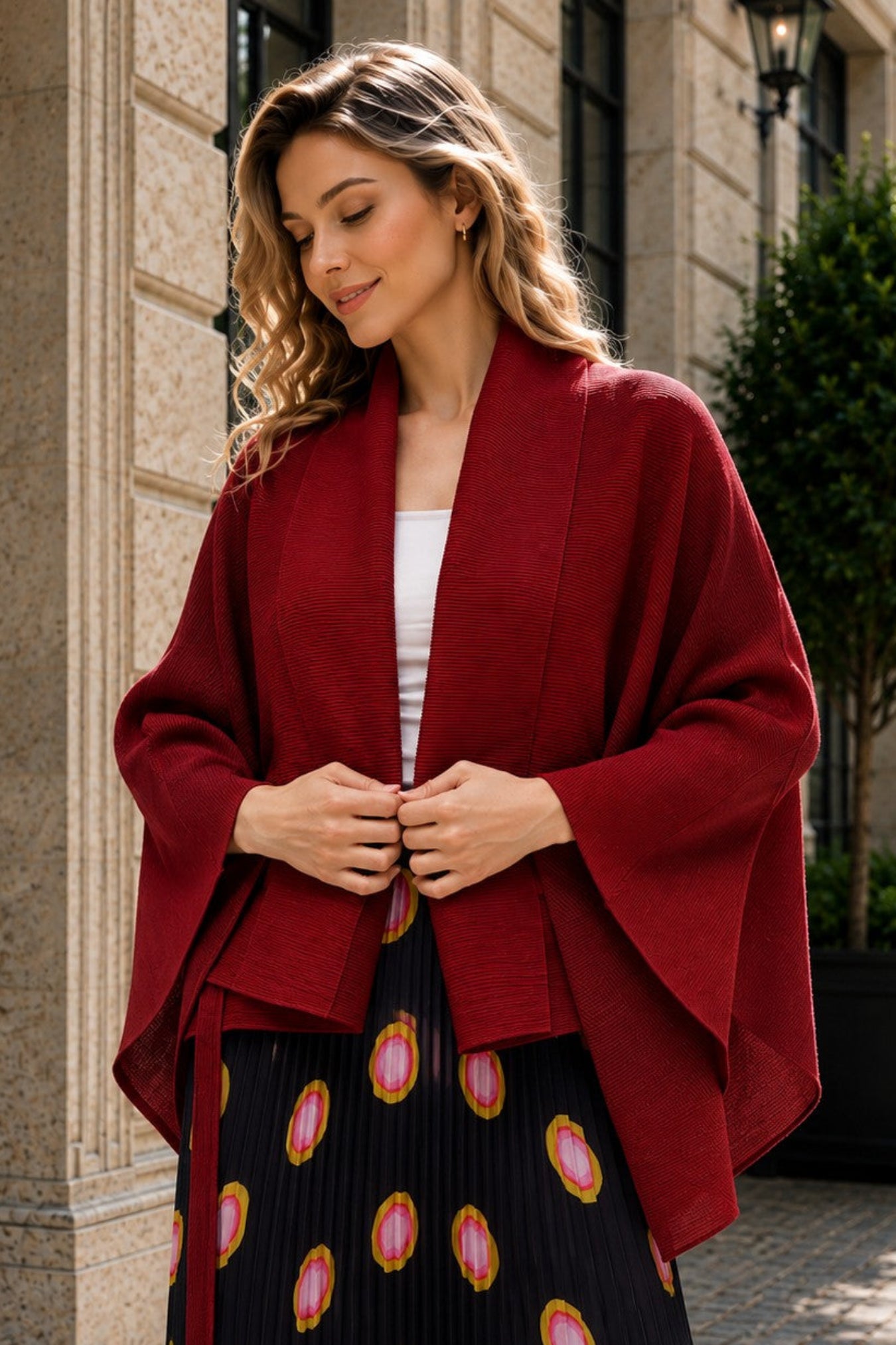 A woman wearing a deep burgundy open-front shawl over a white top and a black pleated skirt with pink and gold circular patterns, standing outdoors in front of a stone building.