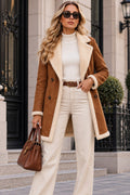 A woman stands outdoors in front of a stone building, wearing a brown suede-like coat with a cream sherpa lining over a white turtleneck, cream wide-leg pants, a brown belt, sunglasses, and carrying a brown handbag.