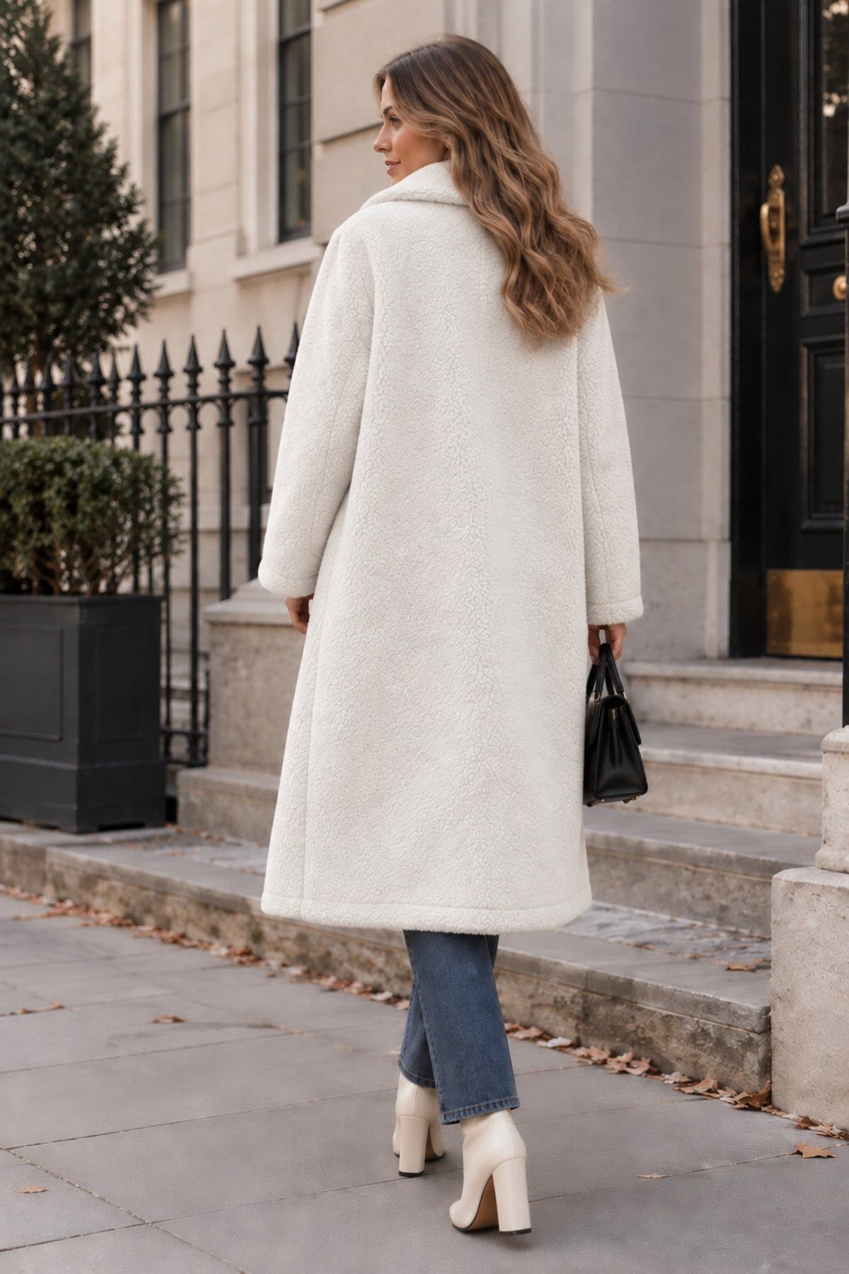 A woman wearing a long cream-colored sherpa teddy coat, blue jeans, and cream ankle boots, standing on a city sidewalk in front of a building entrance.