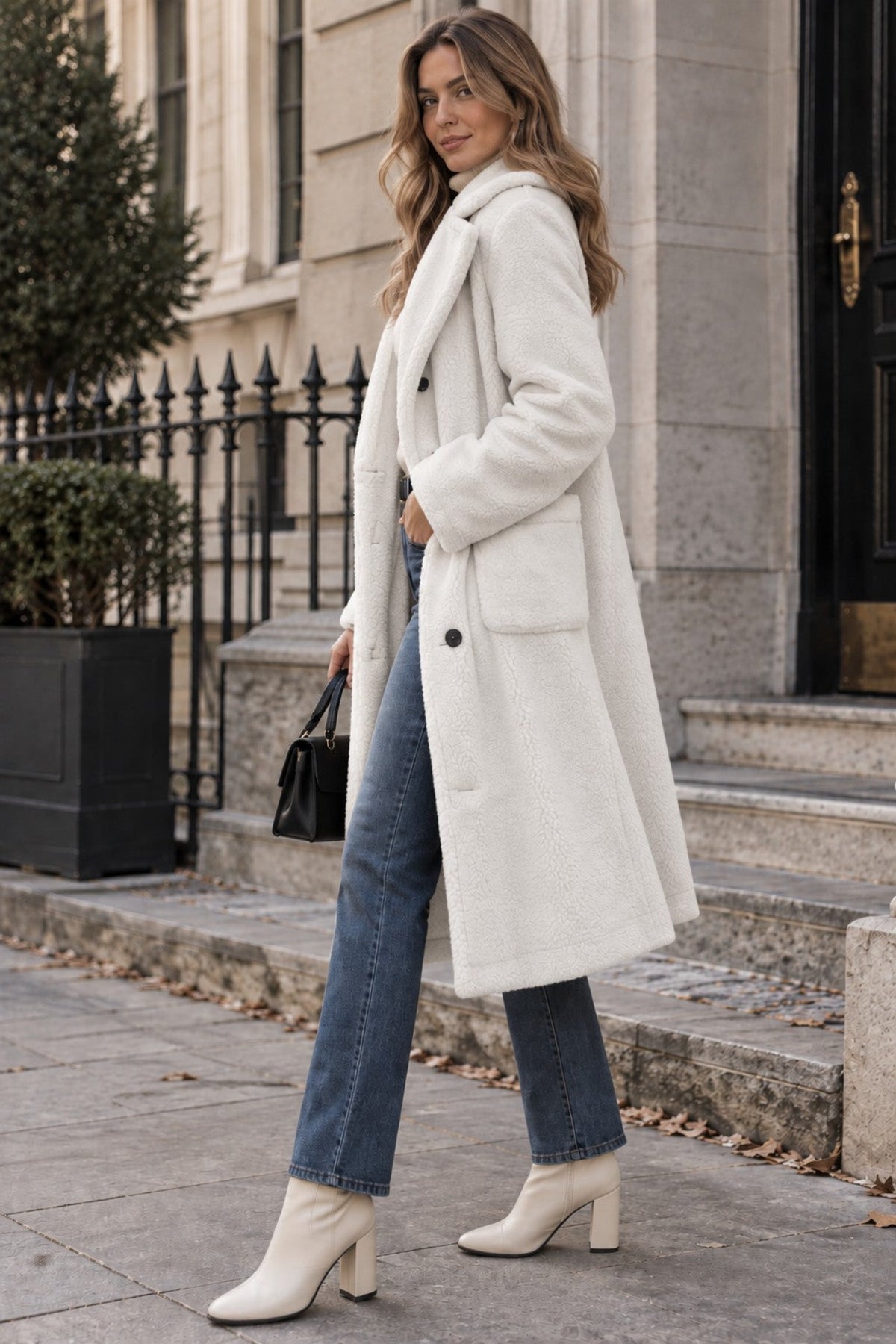 A woman stands on stone steps wearing a long cream-colored faux fur coat, blue jeans, and cream ankle boots, holding a small black handbag.