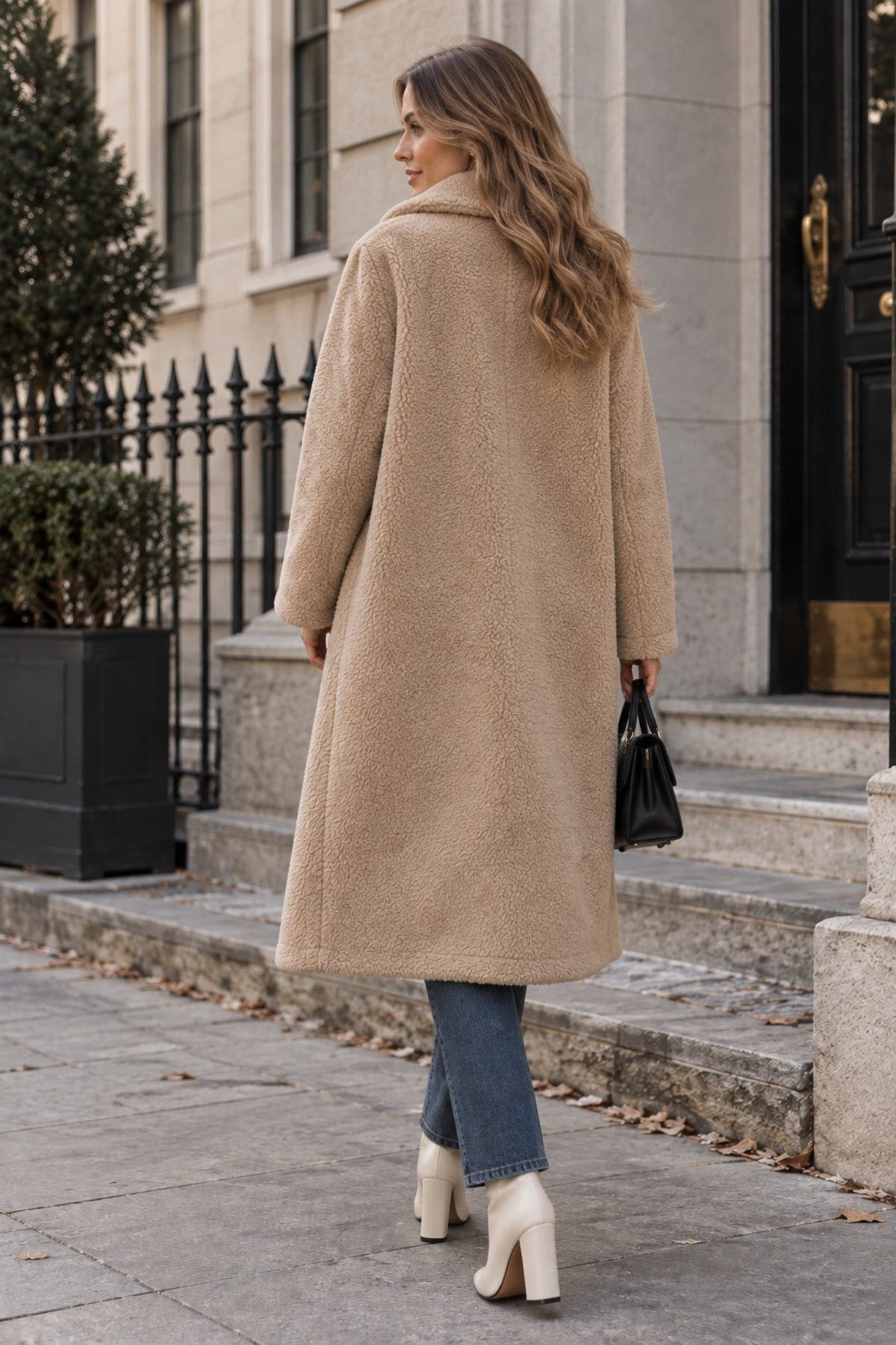 A woman wearing a long beige teddy coat, blue jeans, and cream ankle boots stands on stone steps holding a black handbag.