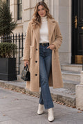 A woman stands on a city sidewalk wearing a long camel teddy coat over a white ribbed turtleneck, blue jeans, and white ankle boots, while holding a small black handbag.
