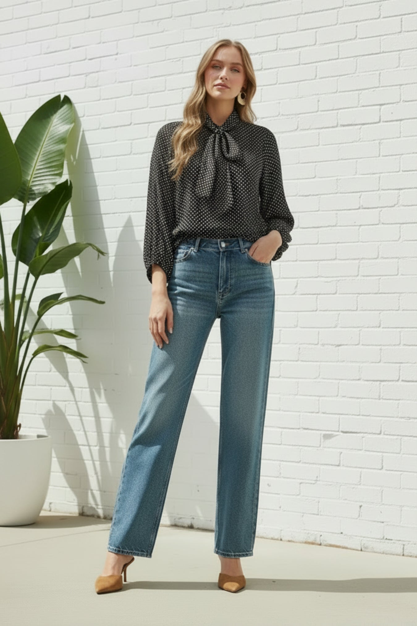 A woman modeling a black and white micro-patterned blouse with a tie-neck detail, paired with blue jeans and brown heels, against a white background next to a plant.