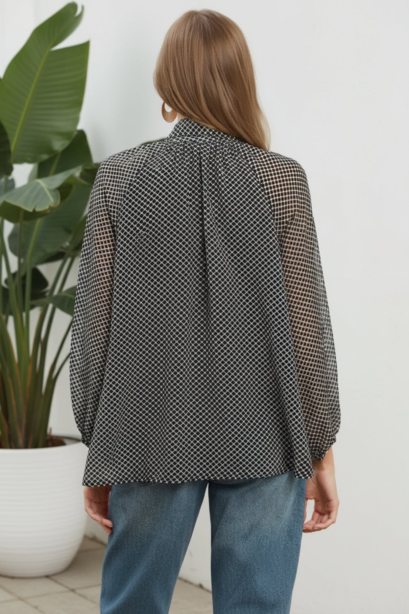 A woman shown from the back wearing a black and white micro-check patterned blouse with sheer long sleeves, paired with blue jeans, against a solid white background with a plant on the side.