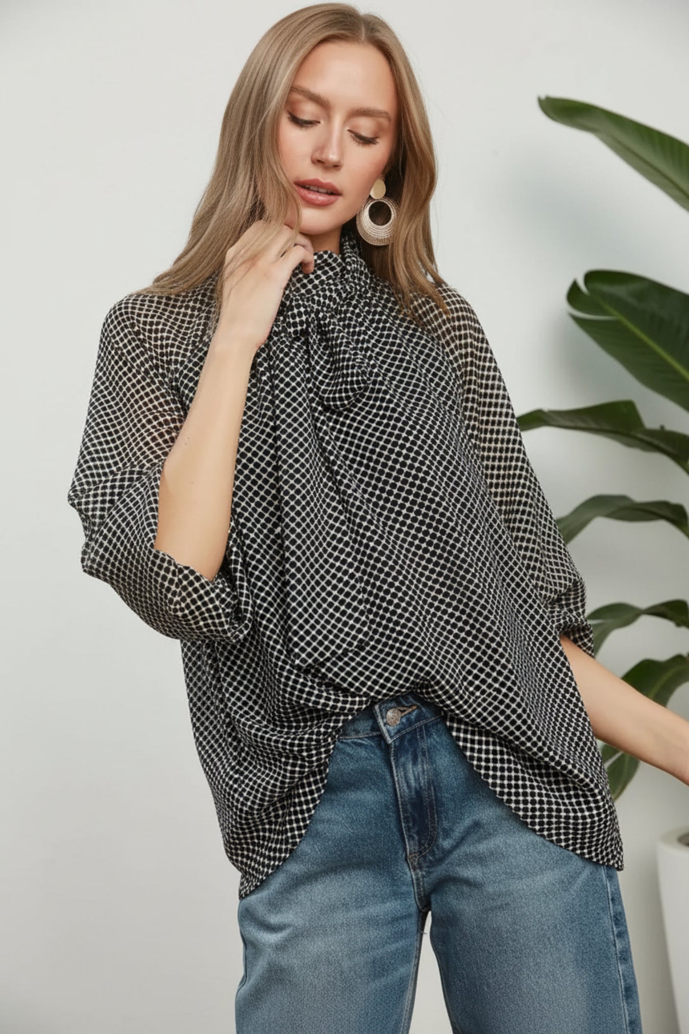 A woman wearing a black and white micro-check patterned blouse with a large ruffled high neck, paired with blue jeans, posed against a white background and a plant.