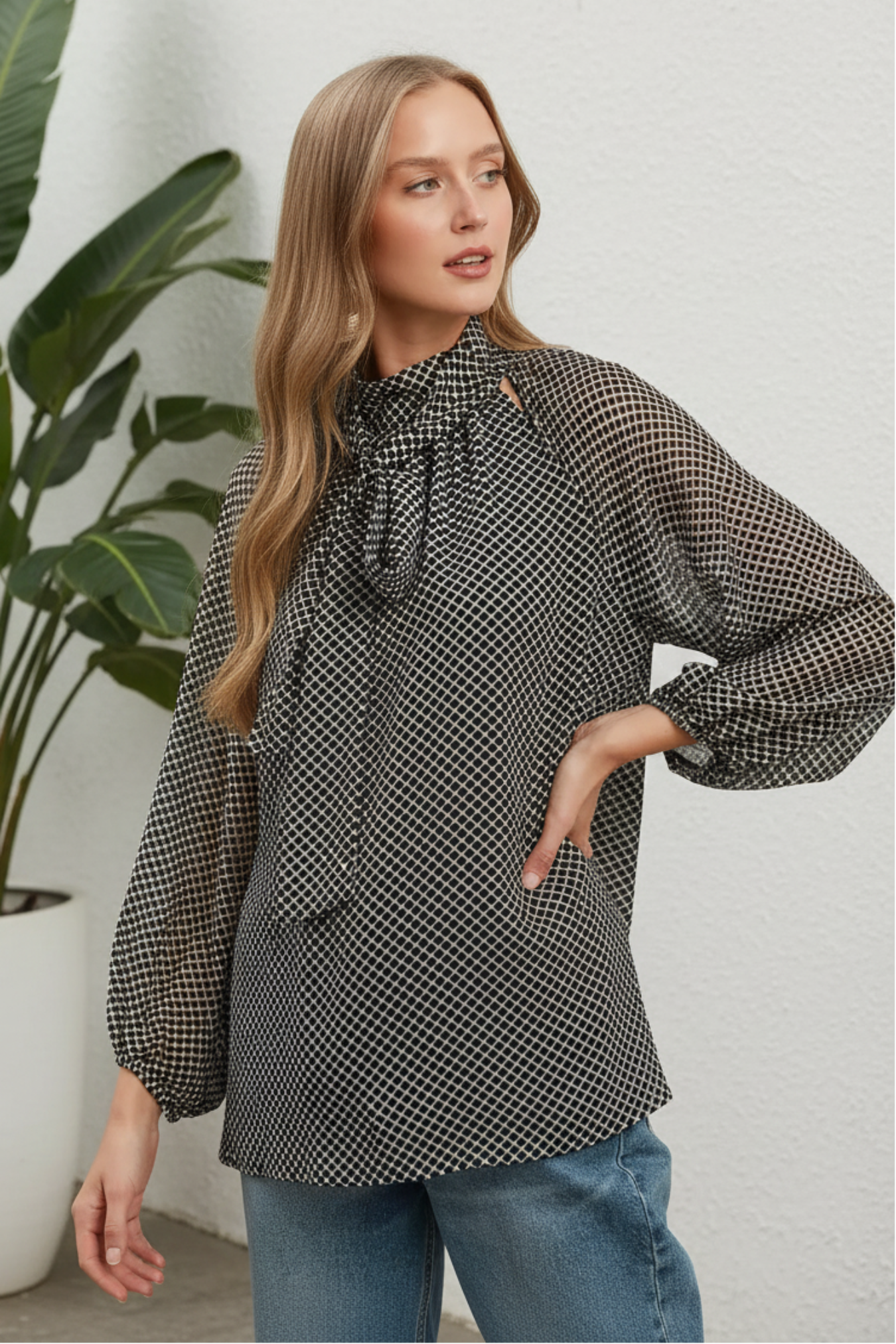 A woman wearing a black and white micro-check patterned blouse with a high neck and large bow detail, paired with blue jeans, set against a white background.