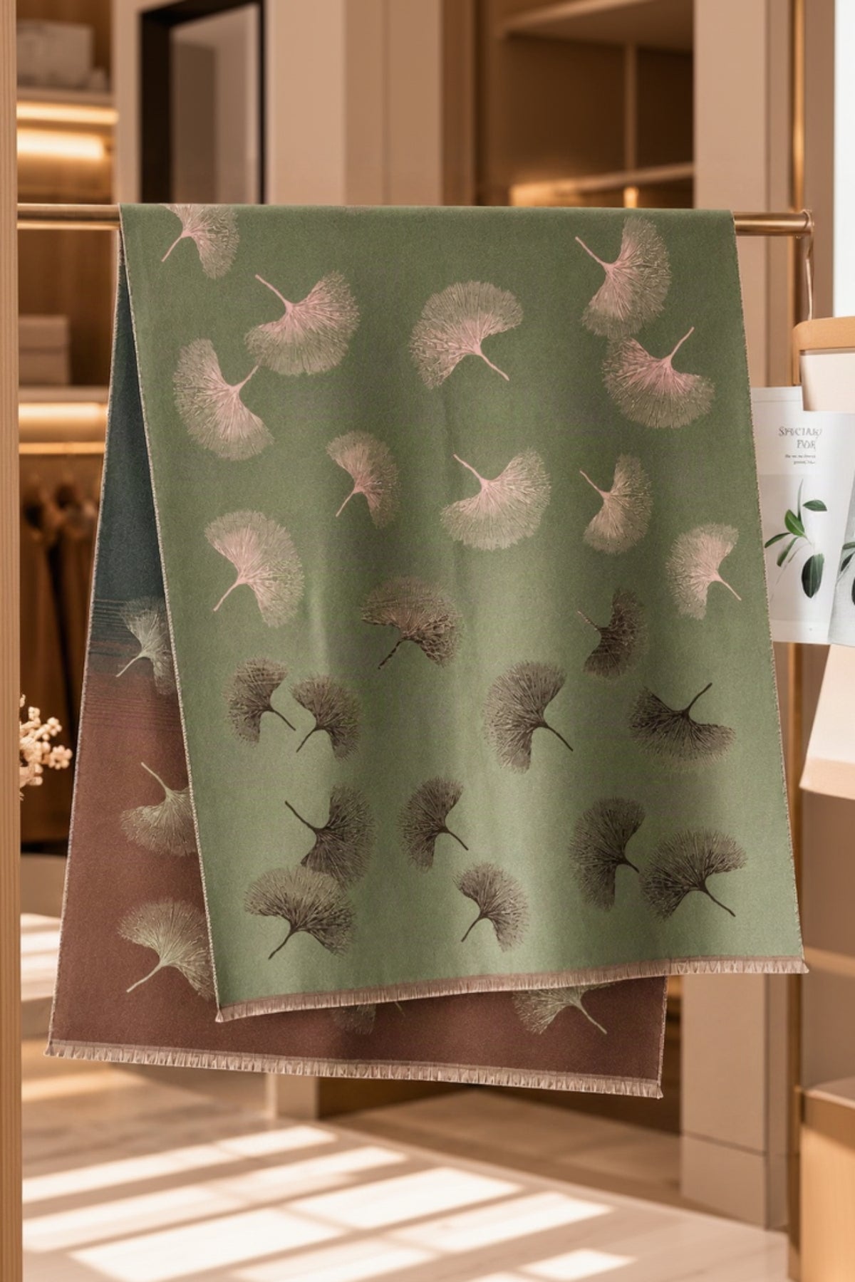 A sage green scarf with a repeating ginkgo leaf pattern in muted taupe and brown tones, draped over a clothing rack in a warm, boutique-style interior.