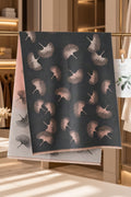 A dark charcoal scarf with a repeating ginkgo leaf pattern in muted pinkish-brown tones, draped over a light gray scarf with a pink ombre gradient. The scene is set in a warm, modern boutique interior with clothing racks and soft lighting.