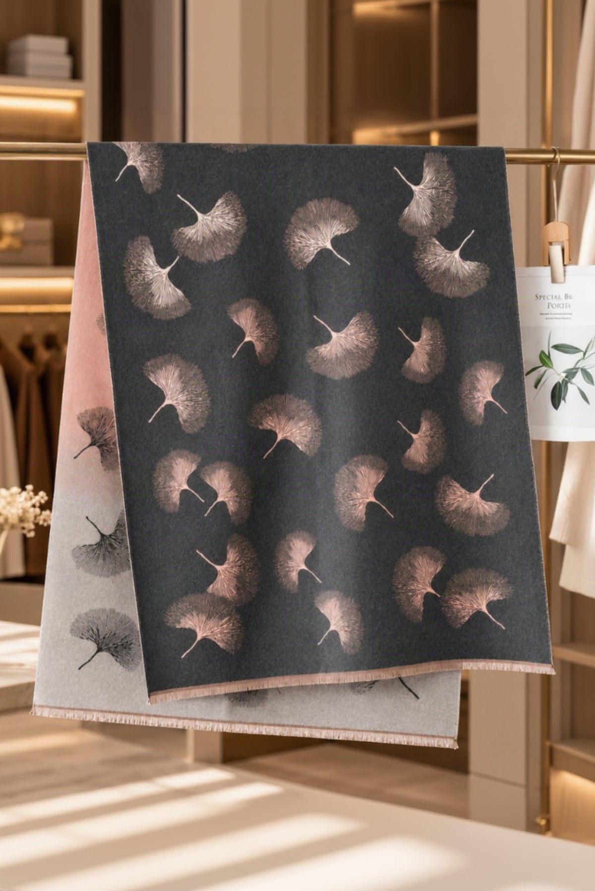 A dark charcoal scarf with a repeating ginkgo leaf pattern in muted pinkish-brown tones, draped over a light gray scarf with a pink ombre gradient. The scene is set in a warm, modern boutique interior with clothing racks and soft lighting.