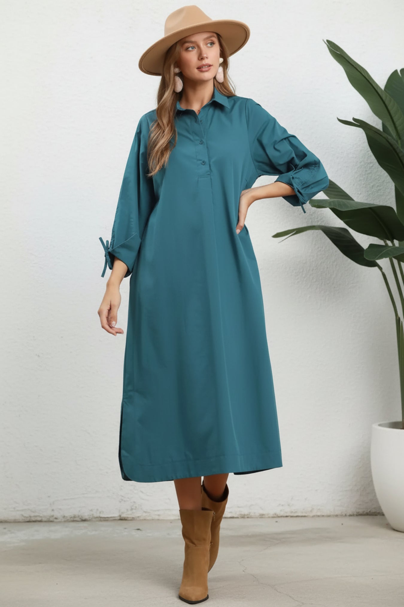 A woman modeling a teal long-sleeve midi shirt dress with a collar and button front, styled with a beige wide-brim hat and beige ankle boots against a white background.