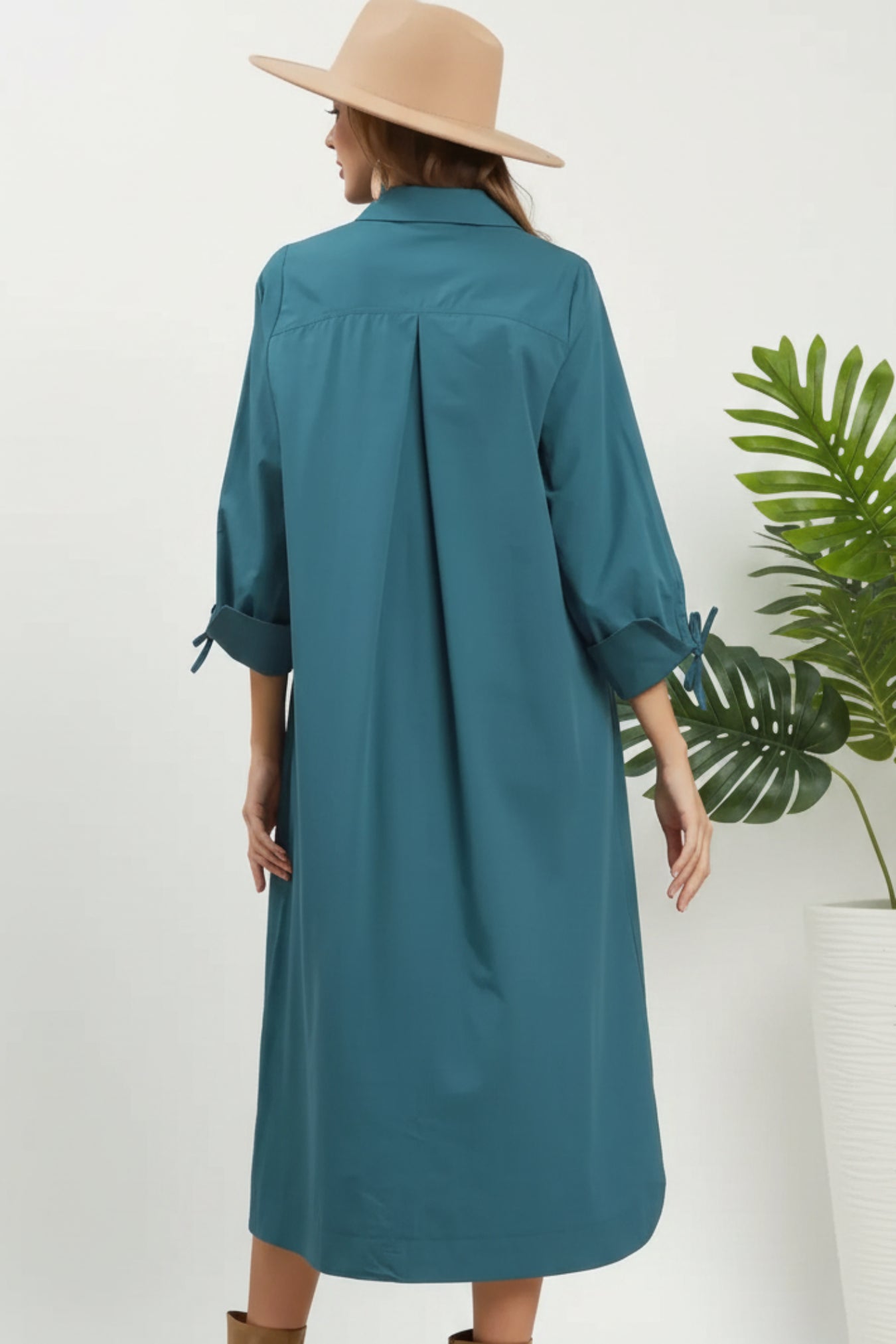 A woman wearing a teal blue long-sleeve shirt dress, shown from the back. She is wearing a beige wide-brim hat and standing next to a green potted plant against a white wall.
