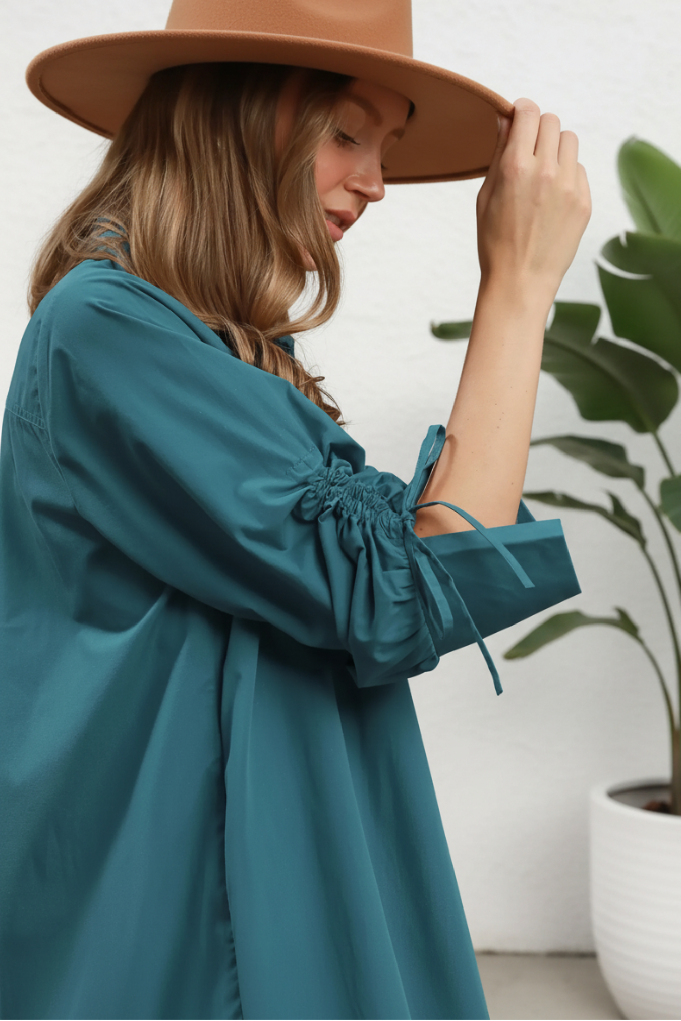A woman wearing a teal long-sleeve blouse with gathered elbow-length sleeves and tie details, shown against a white background and a plant on the side. 