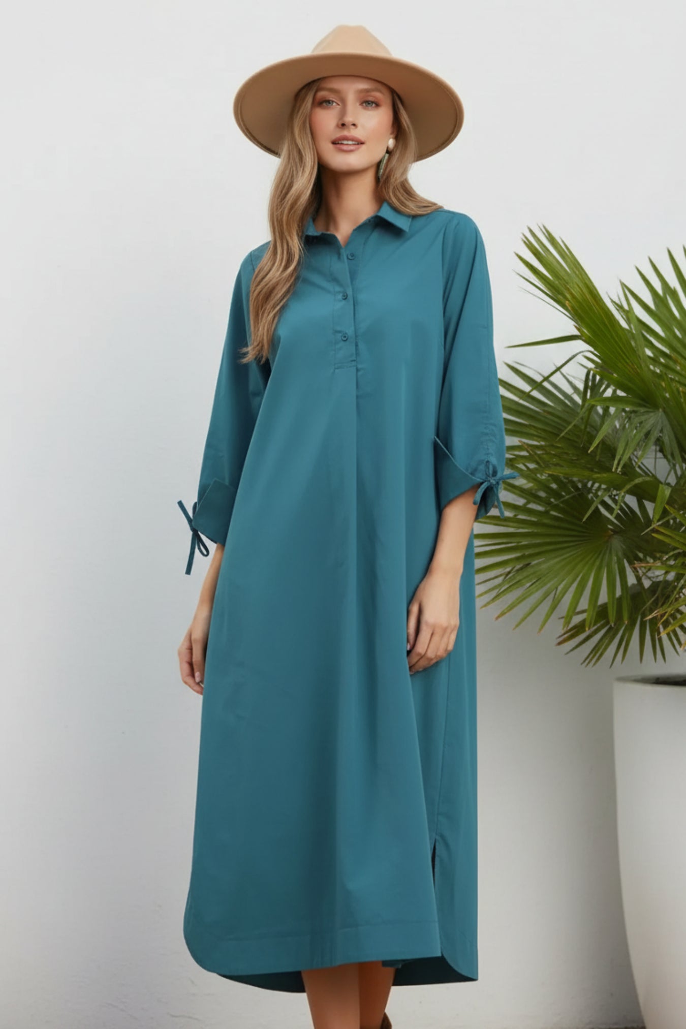 A woman wearing a teal button-front midi shirt dress with three-quarter sleeves and tie details at the cuffs, styled with a wide-brim tan hat against a white background.