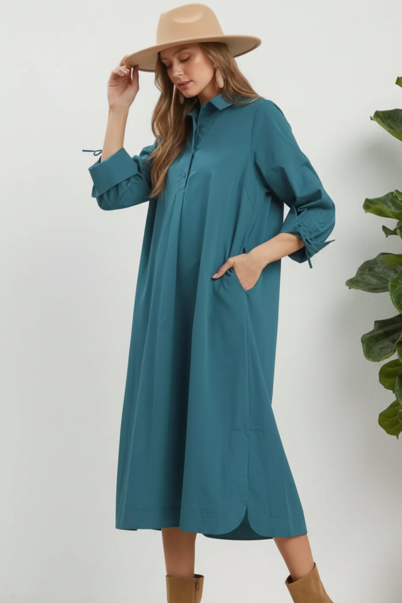 A woman modeling a teal long-sleeve midi shirt dress with a high collar and front pockets, paired with a beige wide-brim hat and tan ankle boots, against a white background.