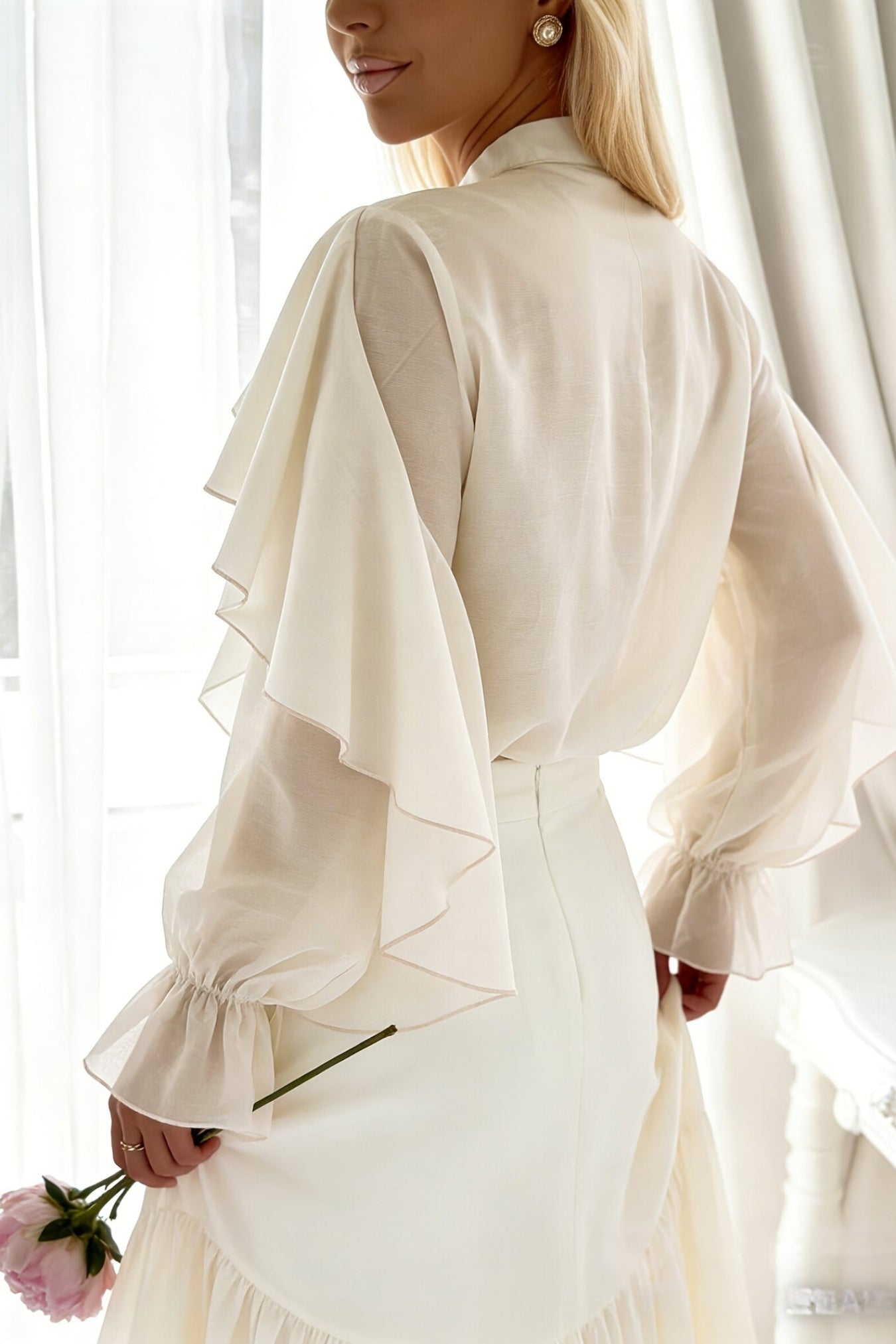 A woman shown from the back wearing a cream-colored high-neck blouse with dramatic layered ruffle sleeves and a matching skirt, holding a pink rose in a bright, softly lit room.