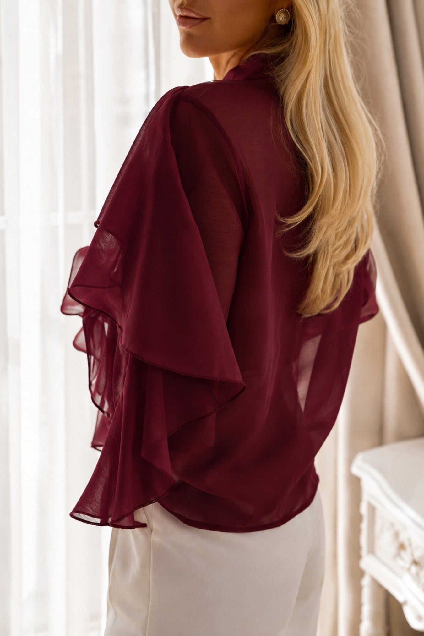 A woman wearing a burgundy sheer ruffled blouse and white pants, shown from a side-back angle in a softly lit indoor setting with curtains and a white table.