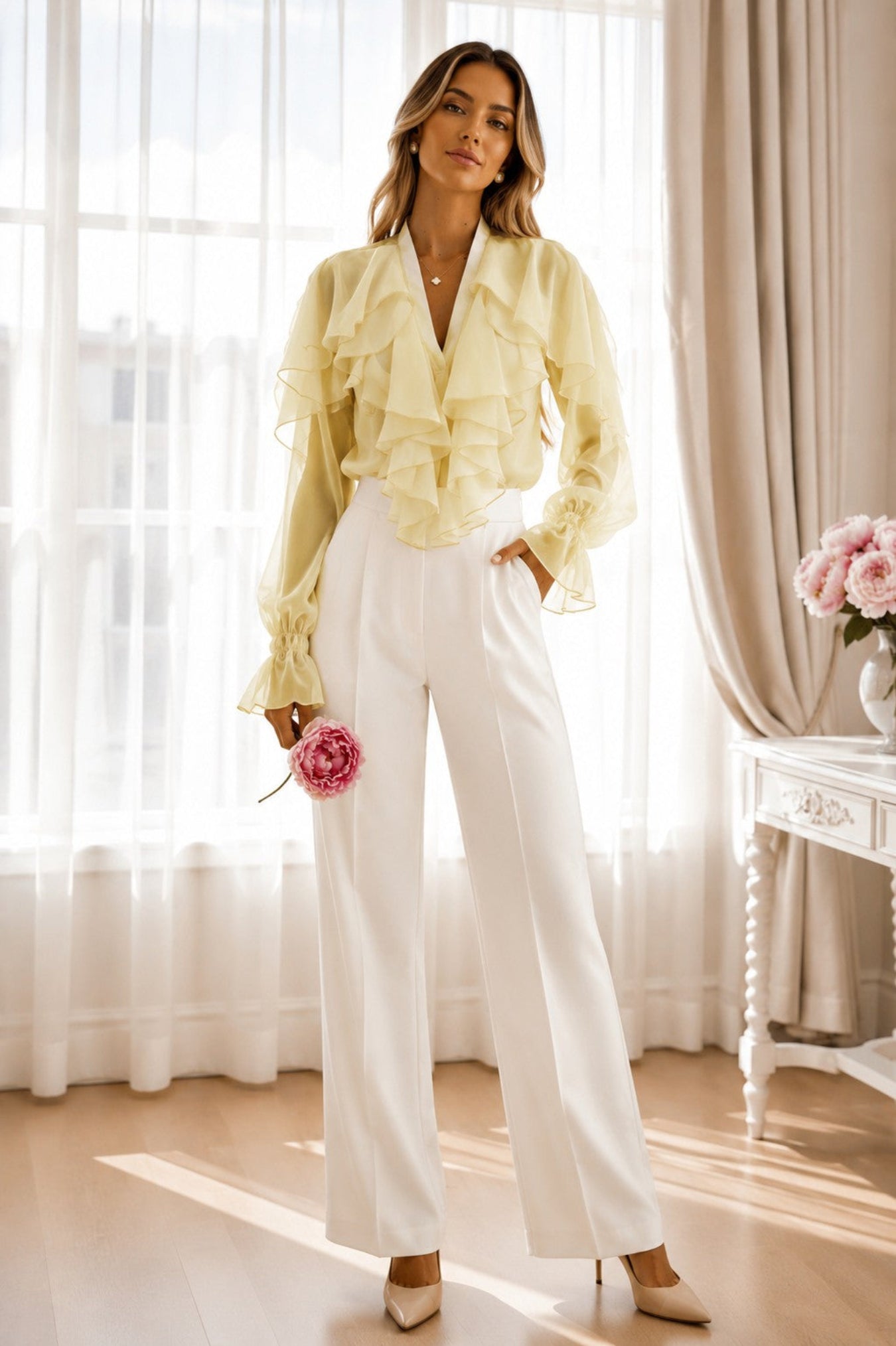 A woman stands in a bright room wearing a pale yellow ruffled blouse with long bell sleeves and white wide-leg trousers, holding a pink flower. She is posed in front of sheer white curtains and a window, with a small table and pink flowers visible on the right.