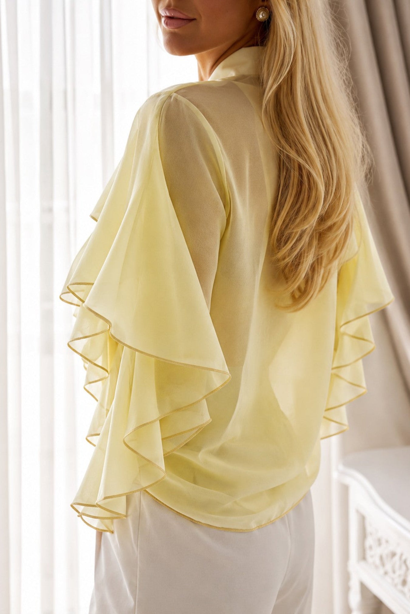 A woman wearing a pale yellow ruffled blouse and white pants, shown from the back in a softly lit indoor setting with curtains and a white table.