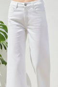 White cropped wide-leg jeans with a ruffled hem, shown on a model wearing beige platform sandals against a white wall with a green plant leaf on the left.