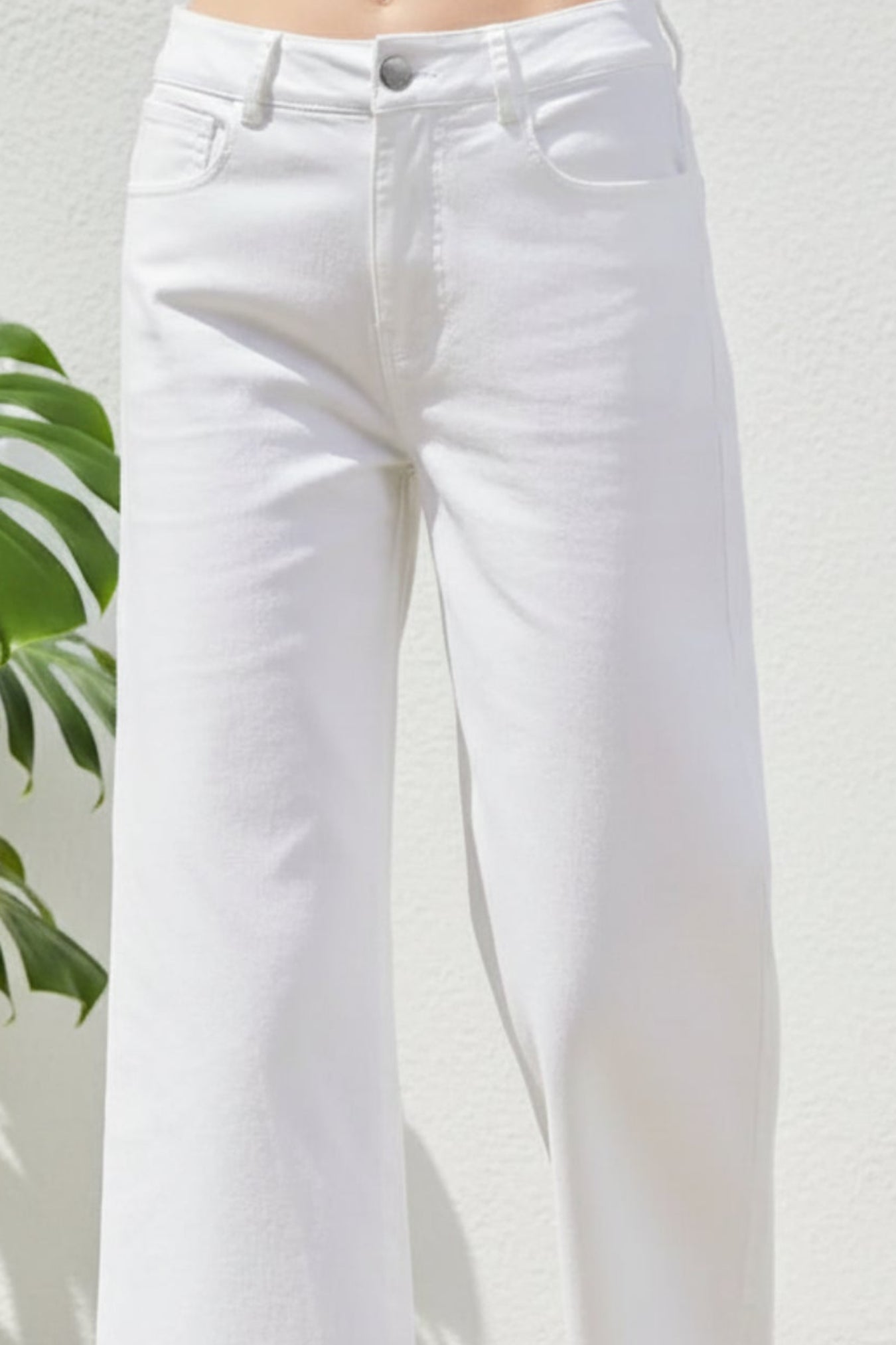 White cropped wide-leg jeans with a ruffled hem, shown on a model wearing beige platform sandals against a white wall with a green plant leaf on the left.