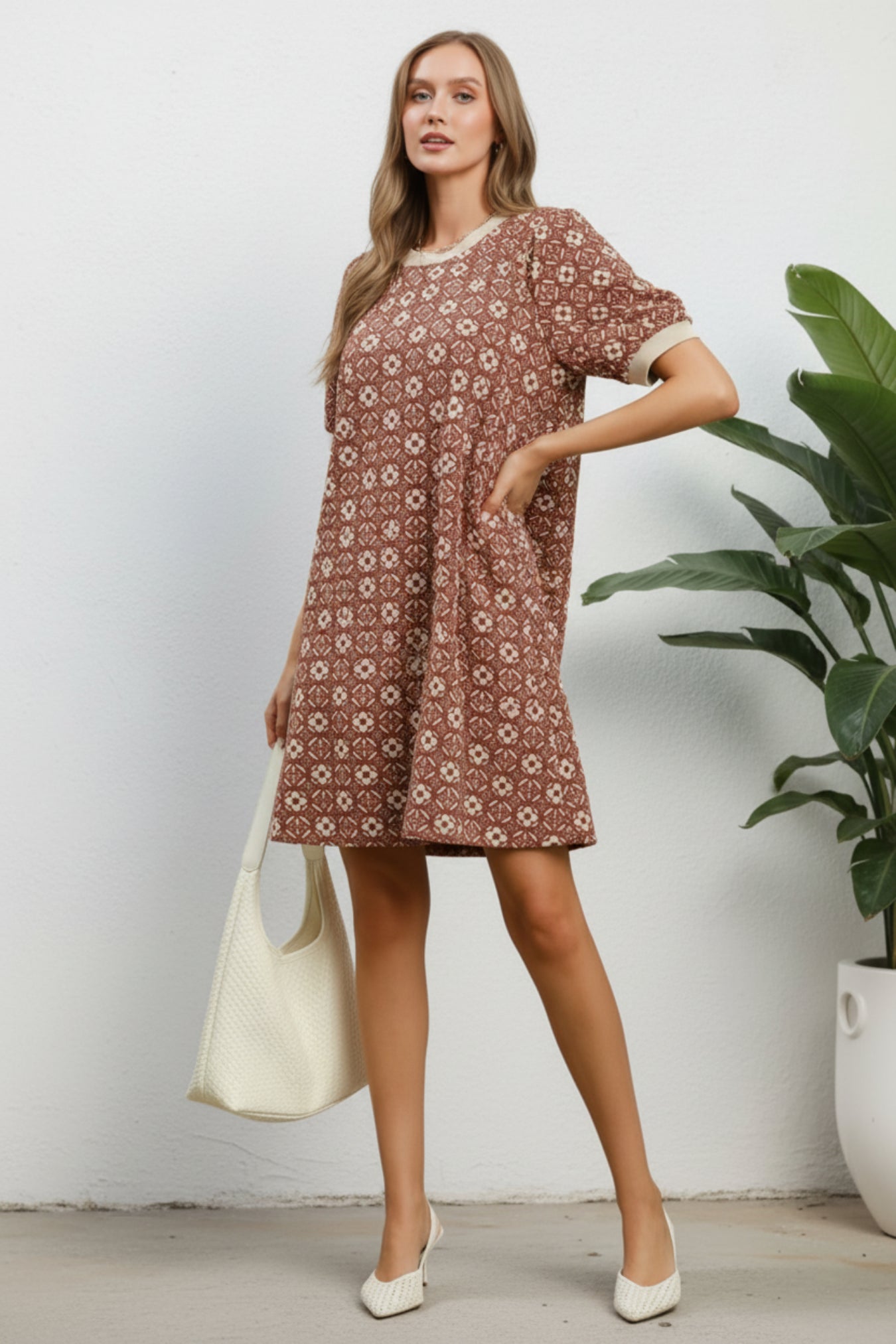 A woman models a short-sleeve brown and beige patterned dress with a crew neck, holding a large cream tote bag and wearing white pointed shoes, set against a white wall with a green potted plant.