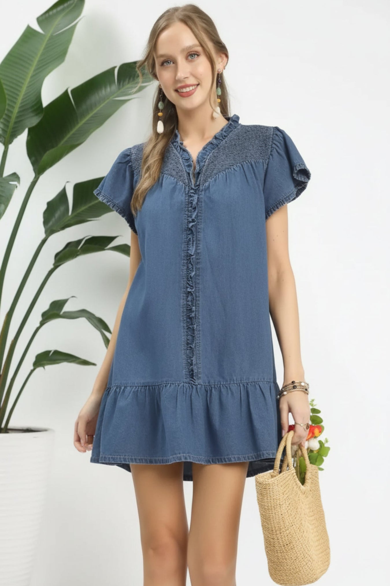 Woman wearing a blue denim dress standing next to a plant indoors.