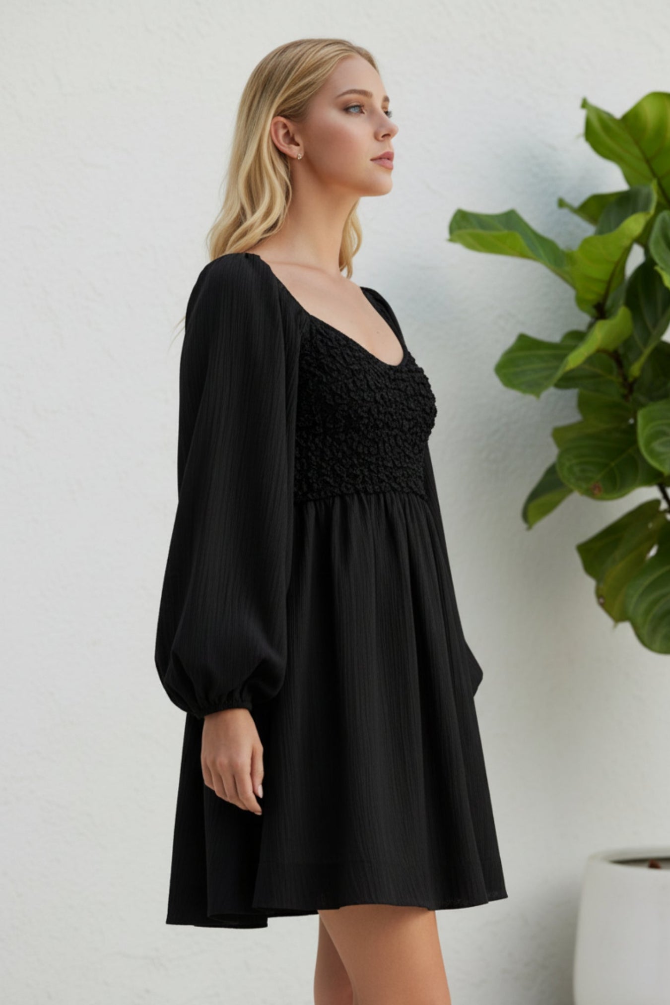 A woman wearing a black mini dress with a smocked bodice and long balloon sleeves, standing in front of a white wall next to a green potted plant.