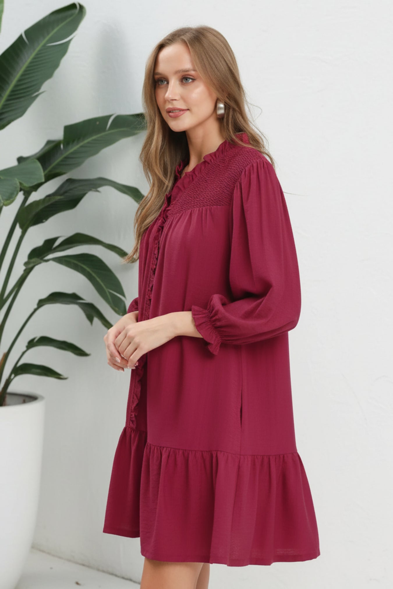 Woman wearing a mulberry dress with lace details on a white background next to a plant