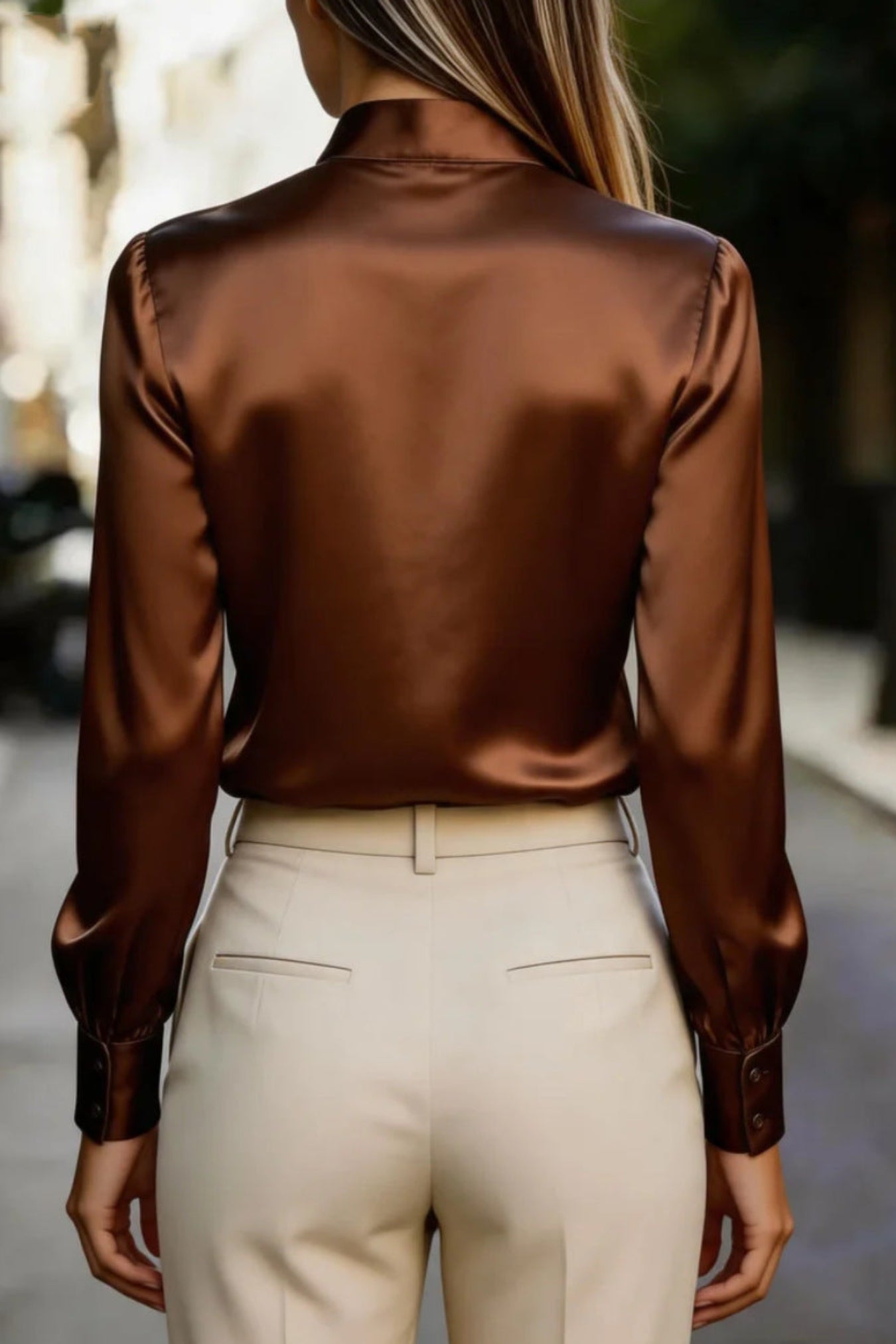 A woman viewed from behind wearing a shiny bronze-brown satin long-sleeve blouse tucked into cream-colored trousers, set against a blurred outdoor street background.