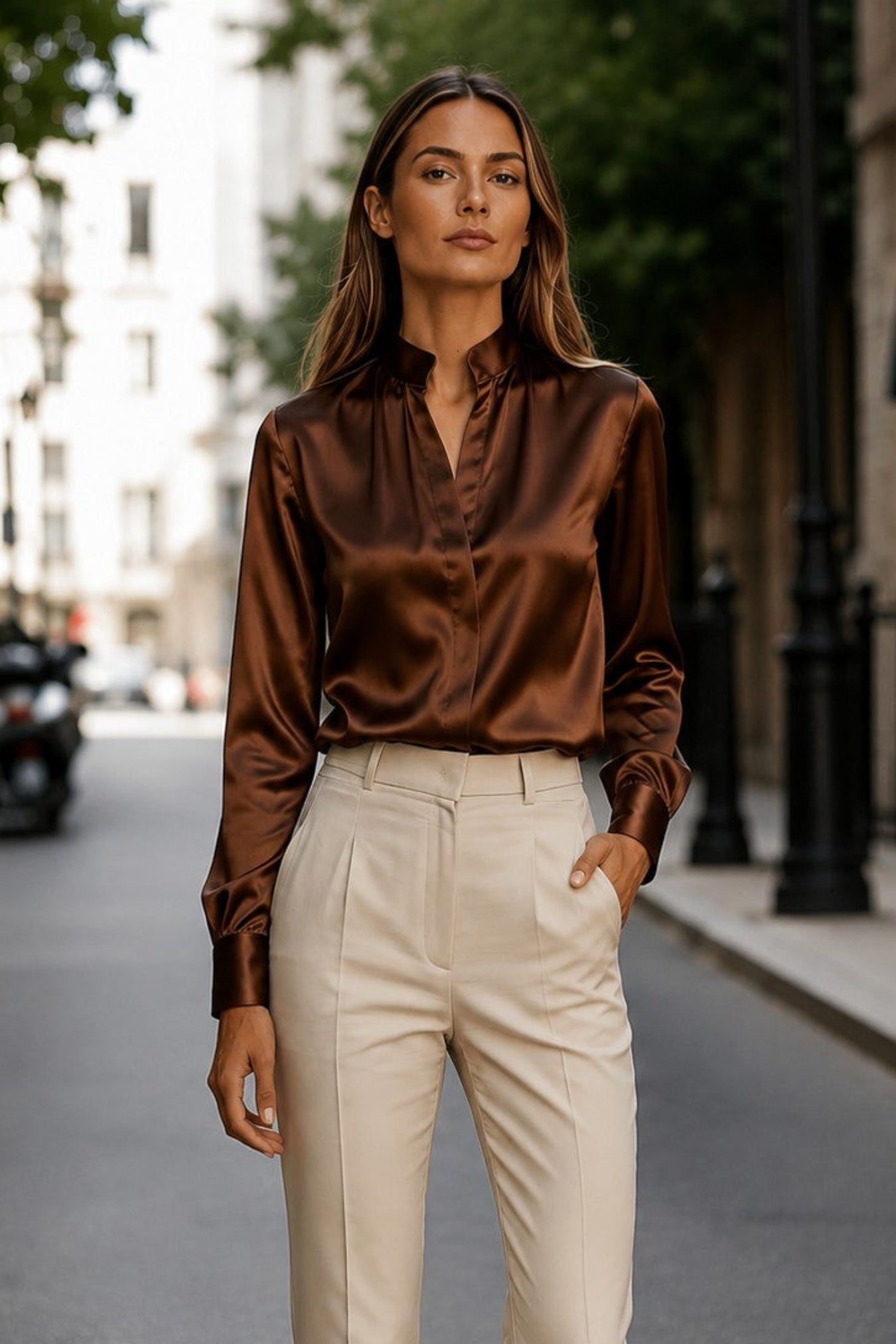 A woman stands on a city street wearing a shiny bronze-brown satin long-sleeve blouse tucked into high-waisted cream trousers, with nude heels.