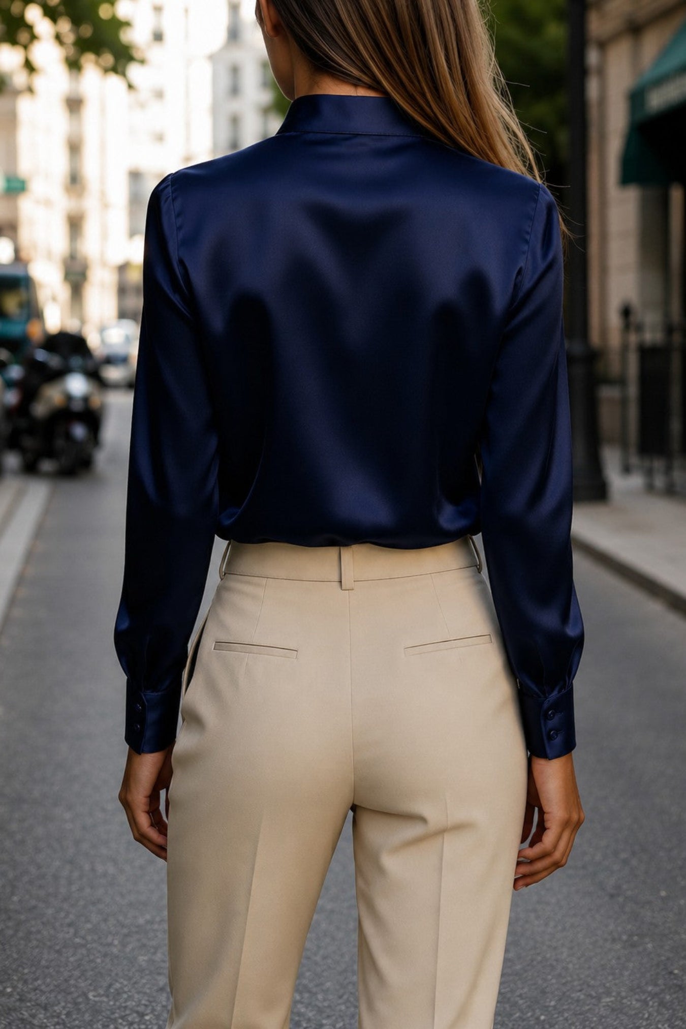 A woman viewed from behind wearing a dark navy satin long-sleeve blouse and beige trousers, standing on a city street.