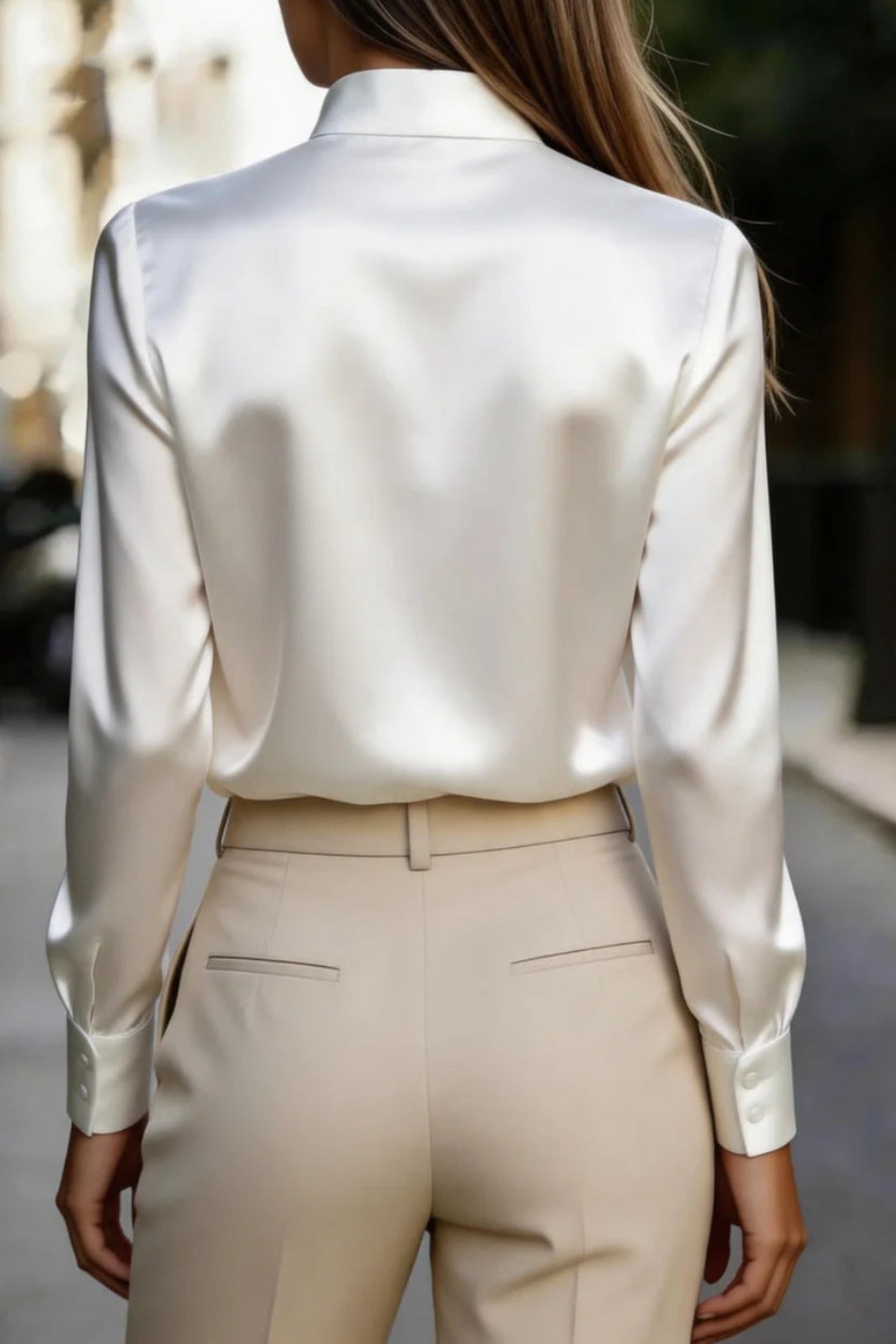 A woman viewed from behind wearing a white satin long-sleeve blouse tucked into light beige trousers, set against a blurred outdoor street background.