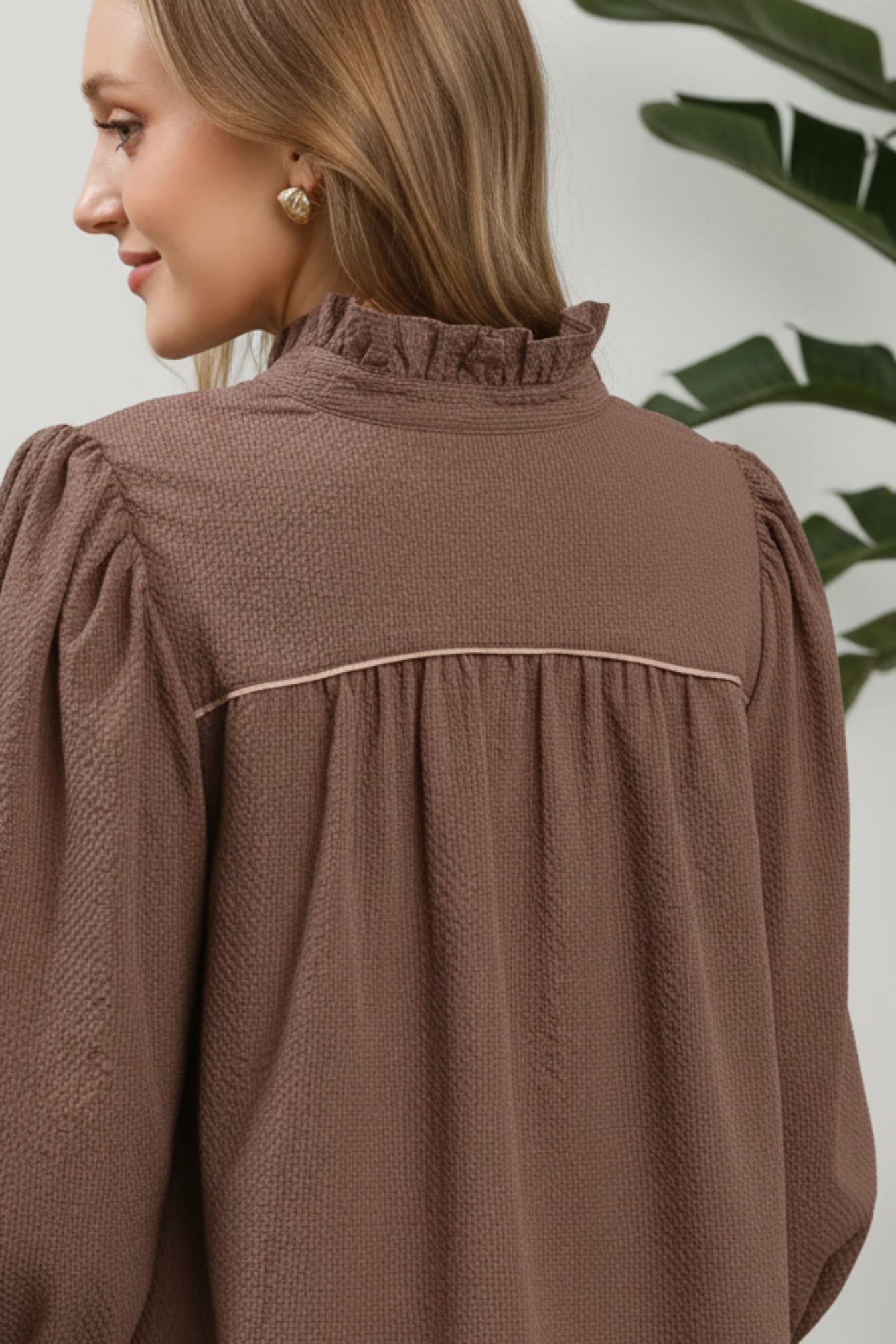 A close-up of a woman wearing a textured chocolate brown blouse with a ruffled mock neck and a thin horizontal seam across the upper back, shown against a white background.