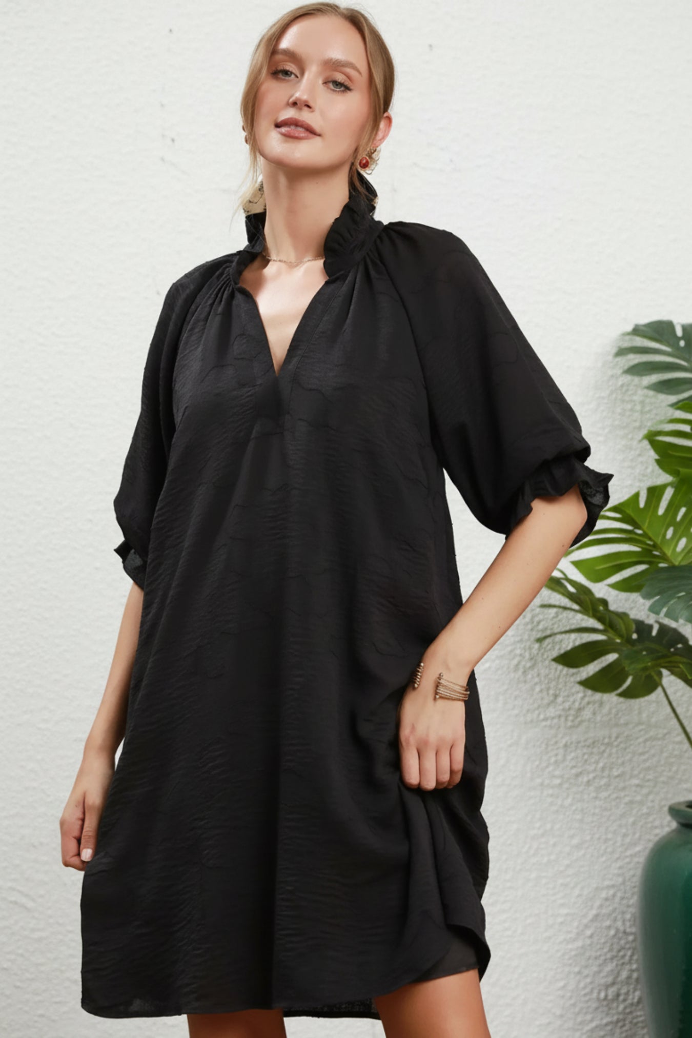 A woman modeling a loose black short-sleeve tunic dress with a gathered high neckline and ruffled sleeves, posed against a white background.