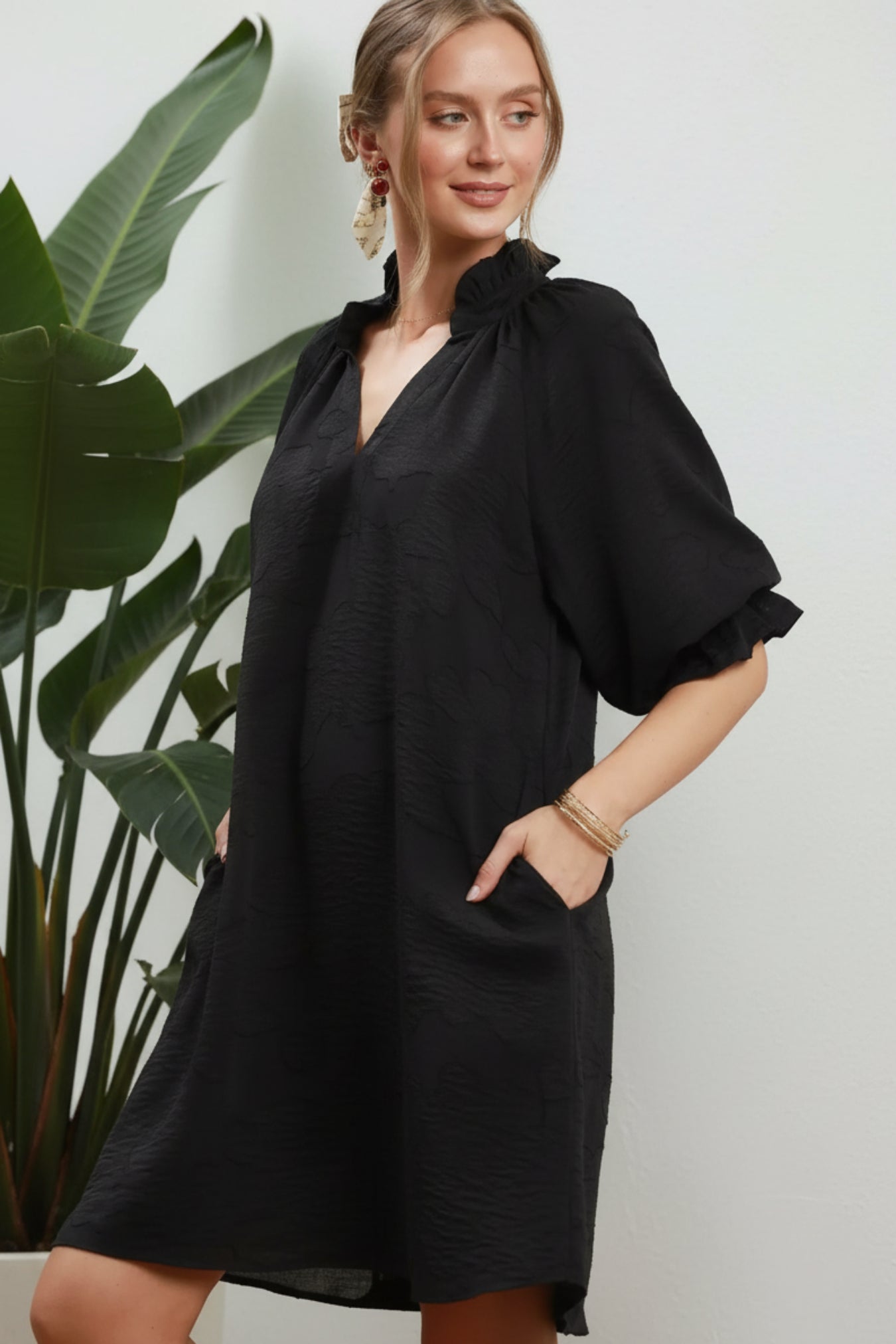 A woman modeling a loose black short-sleeve dress with a gathered high neckline and side pockets, posed against a white background.