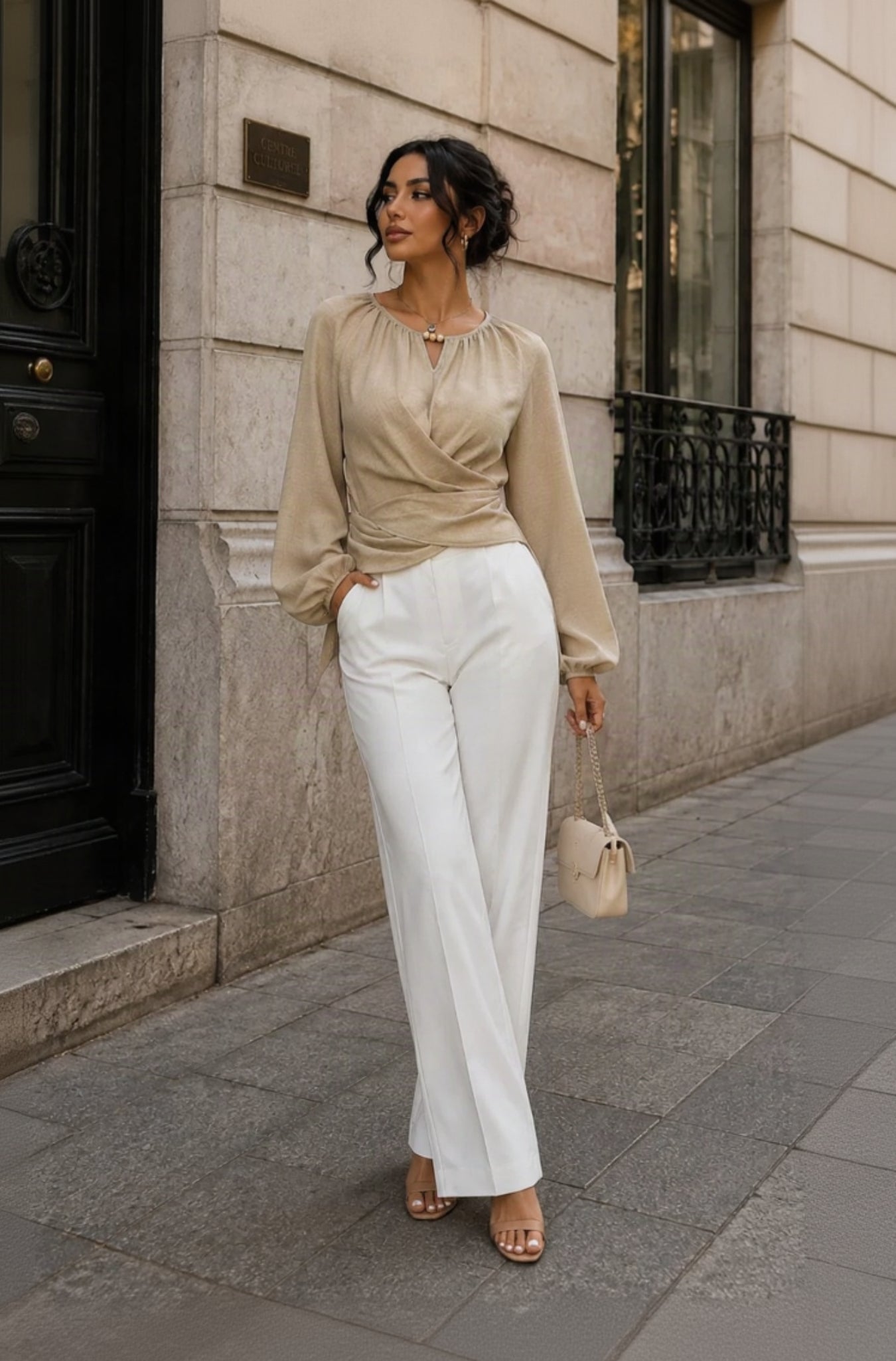 A woman stands on a city sidewalk wearing a beige long-sleeve wrap-style blouse and white wide-leg trousers, with nude heeled sandals and a small beige handbag.
