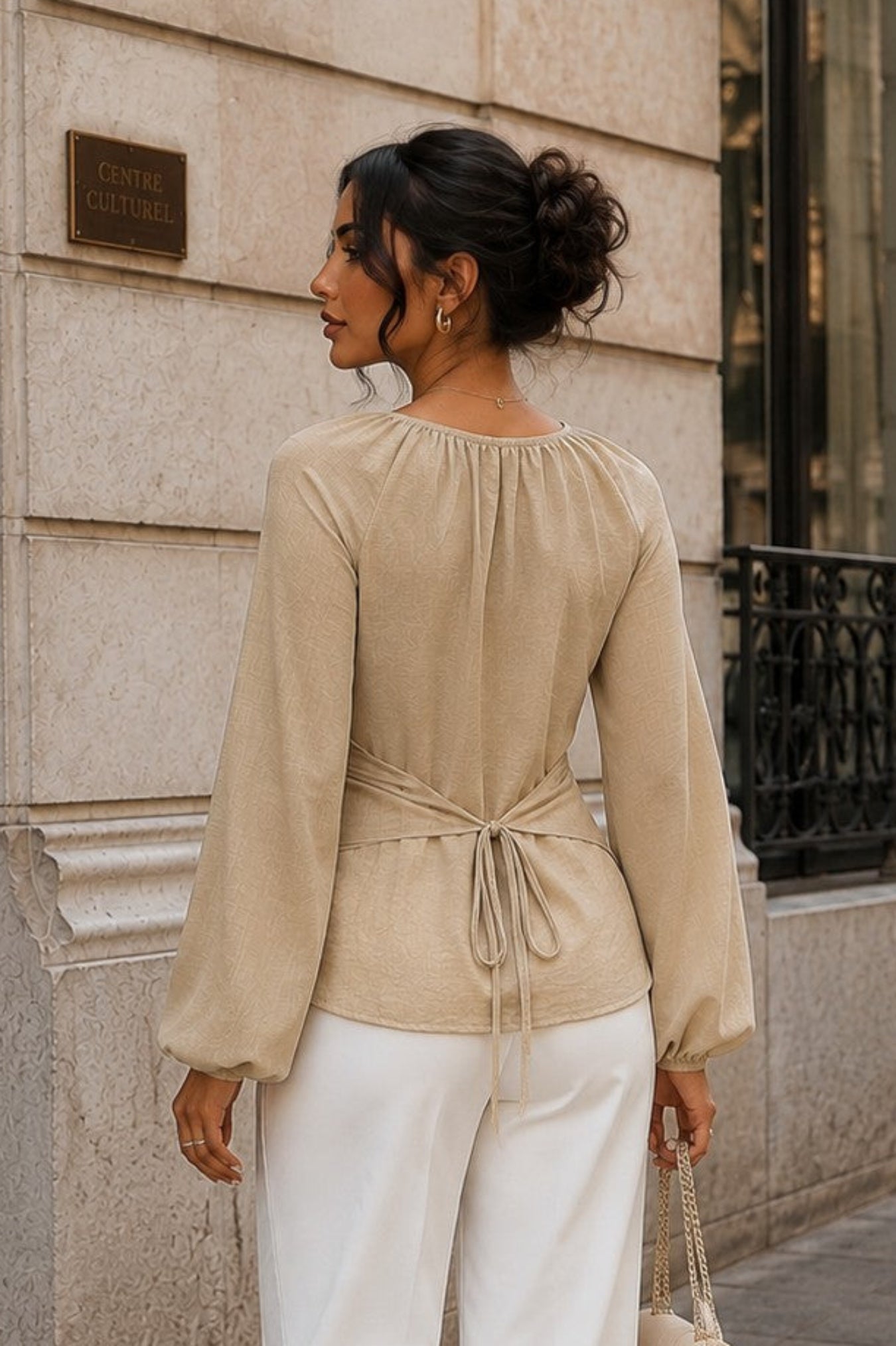 A woman stands on a city sidewalk wearing a beige long-sleeve wrap-style top and white wide-leg pants, holding a small beige handbag.