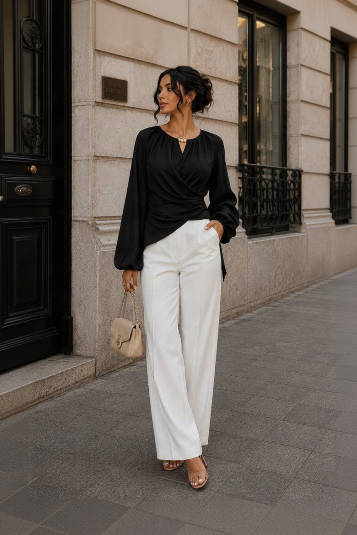 A woman stands on a city sidewalk wearing a black long-sleeve wrap-style blouse and white wide-leg trousers, holding a small beige handbag. The background shows a stone building facade with windows and a dark door.