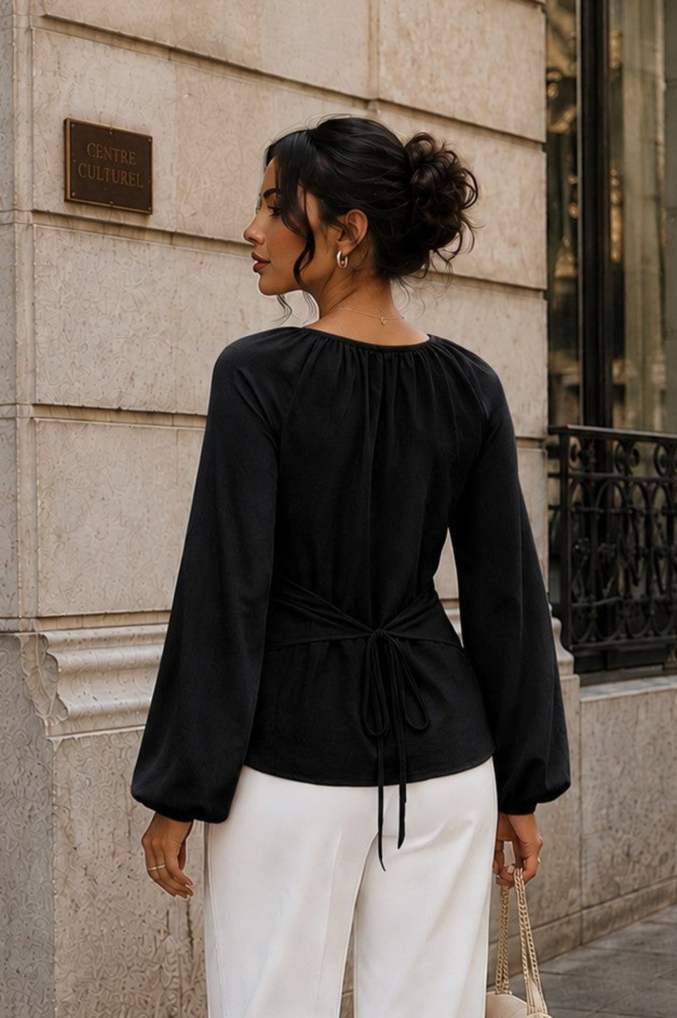 A woman stands on a city sidewalk wearing a black long-sleeve top with a gathered waist and white wide-leg pants, holding a small beige handbag.