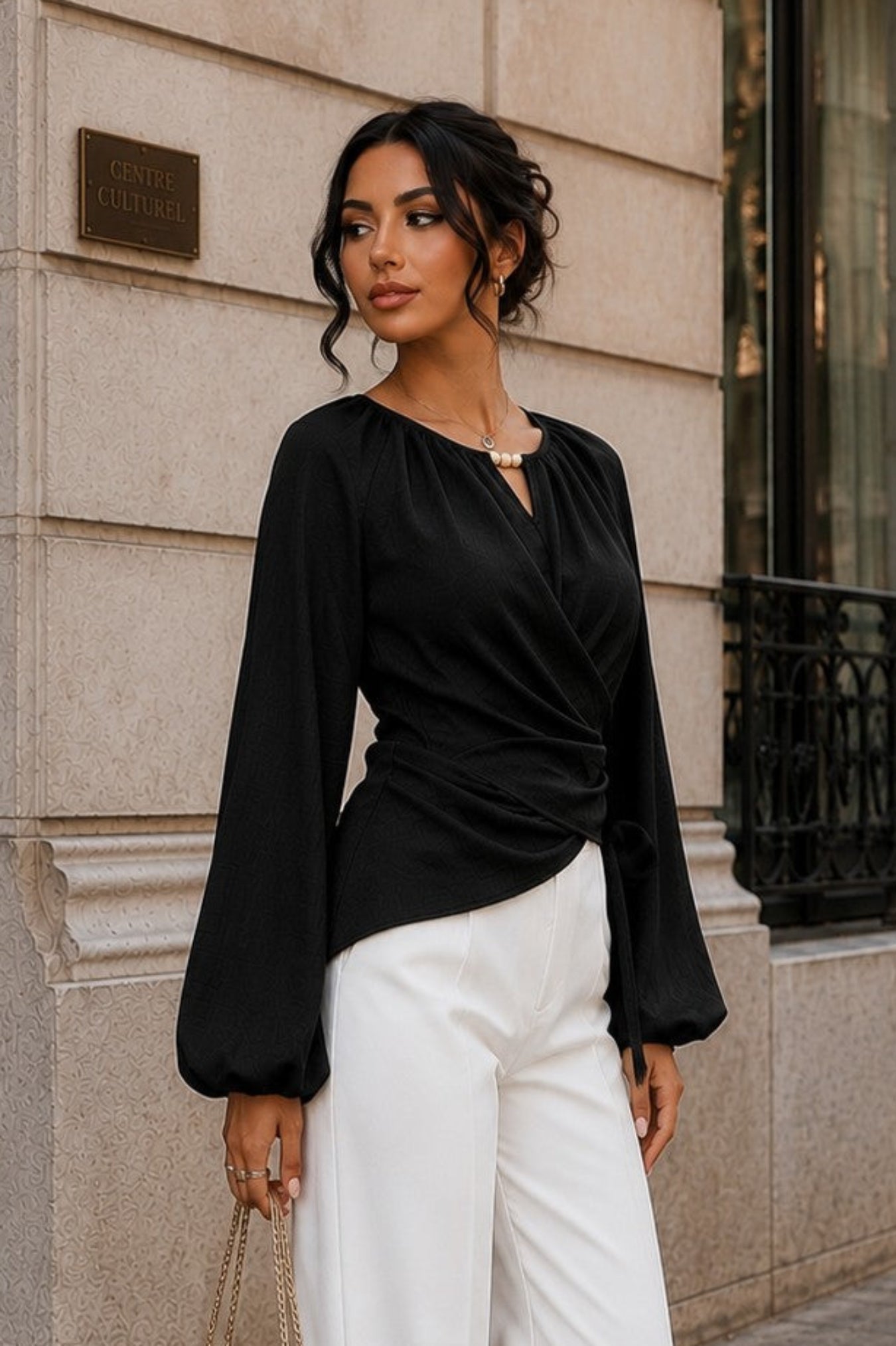 A woman stands on a city sidewalk wearing a black long-sleeve wrap-style blouse with dramatic bell sleeves and white wide-leg trousers, holding a beige handbag.