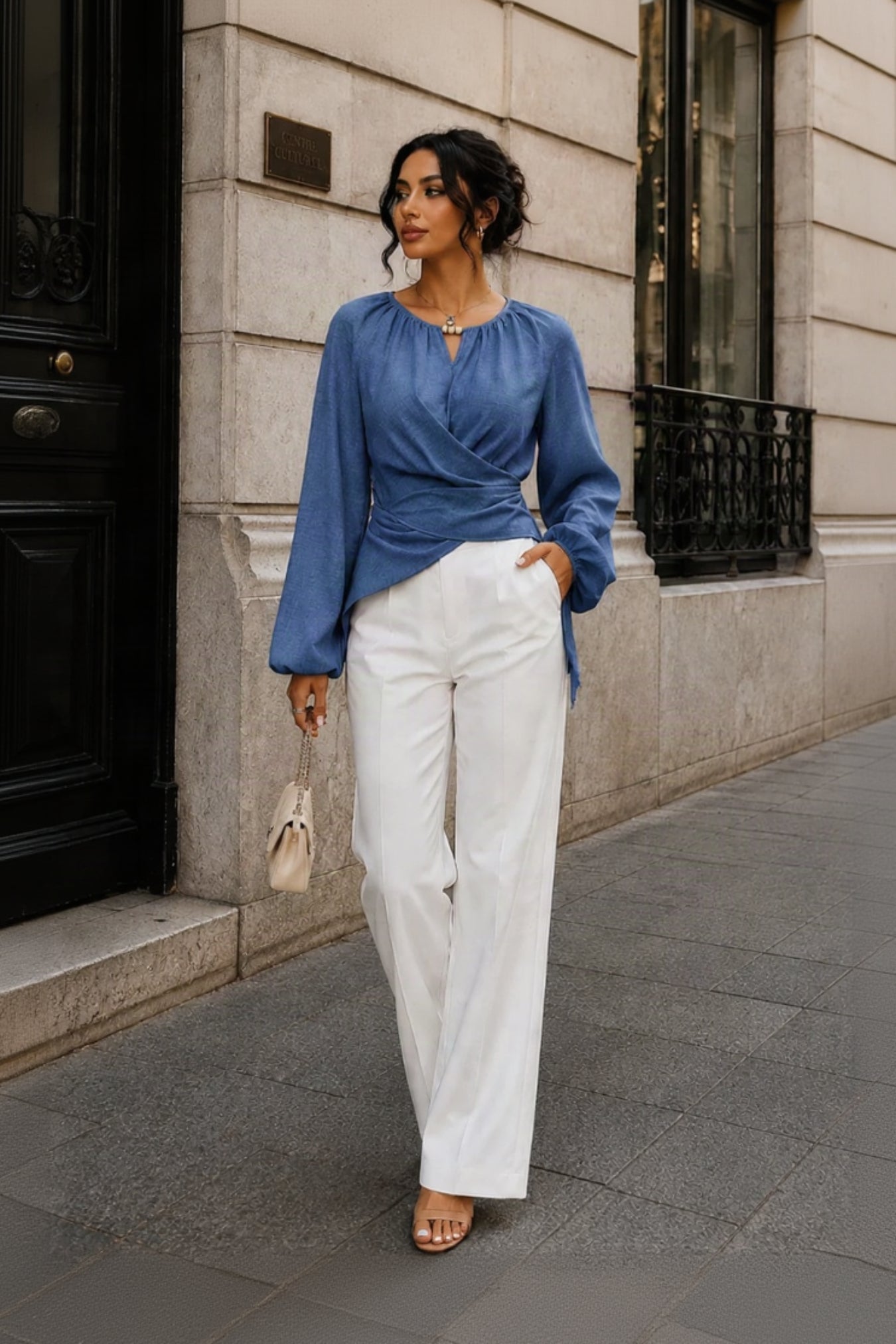A woman stands on a city sidewalk wearing a blue long-sleeve wrap-style blouse with a keyhole neckline and white wide-leg trousers, holding a small beige handbag.