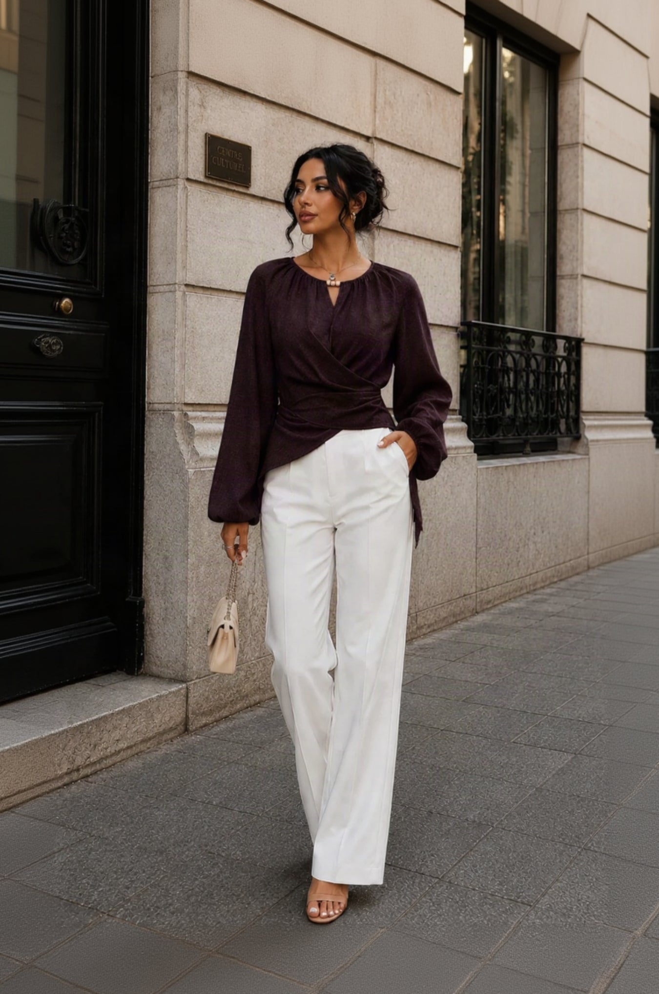 A woman stands on a city sidewalk wearing a dark plum wrap-style long-sleeve blouse and white wide-leg trousers, holding a small beige handbag.